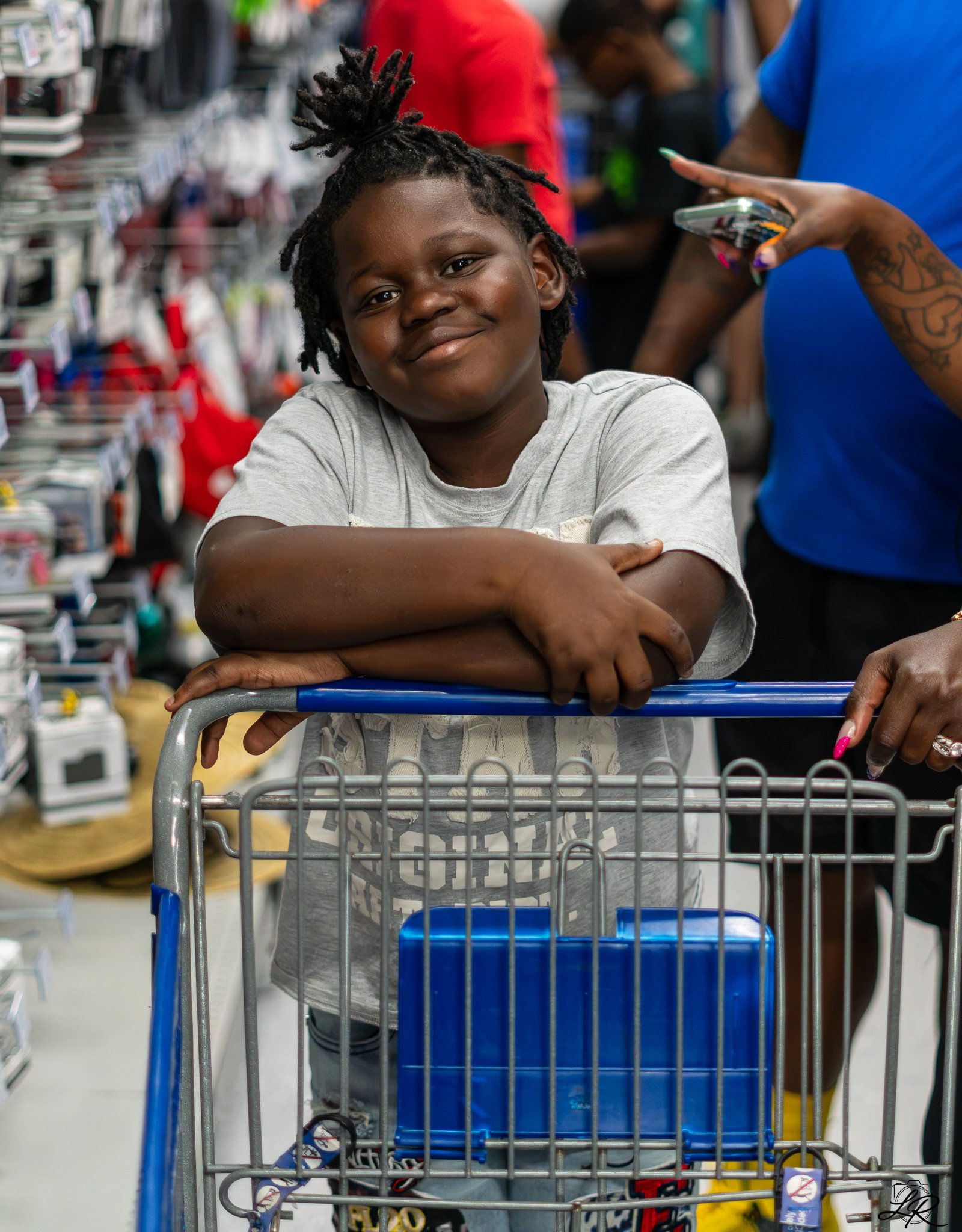 A young boy with dreadlocks leaning on a shopping cart in a store, smiling at the camera, with shelves of household items and other shoppers in the background.