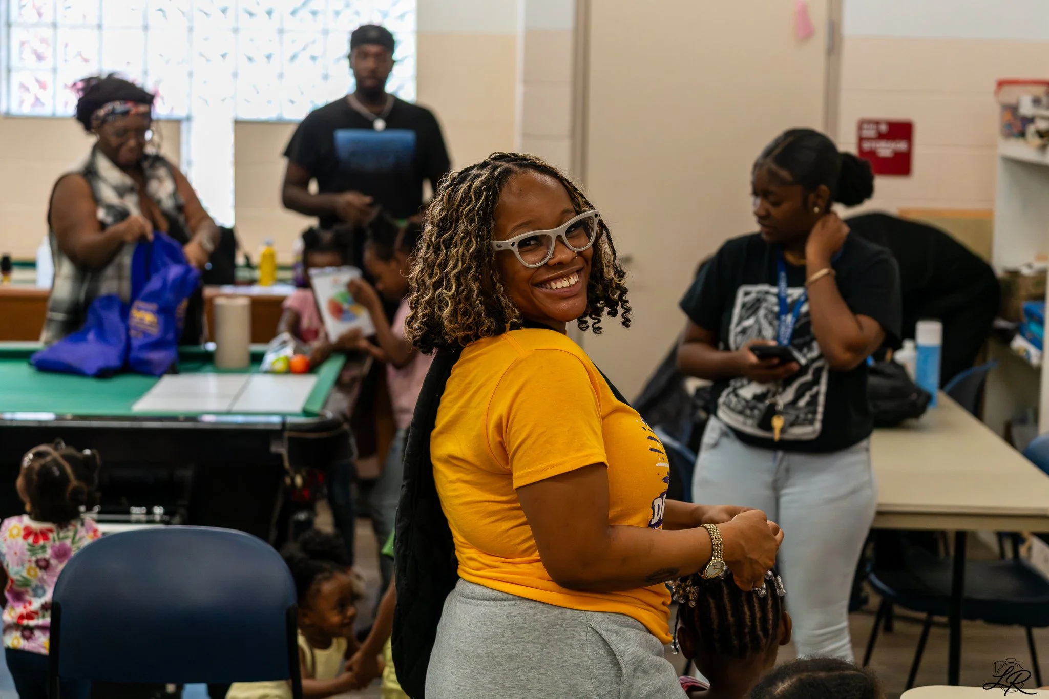 A smiling woman in a yellow t-shirt and glasses looks to her right in a classroom or workshop setting, surrounded by other adults and children.
