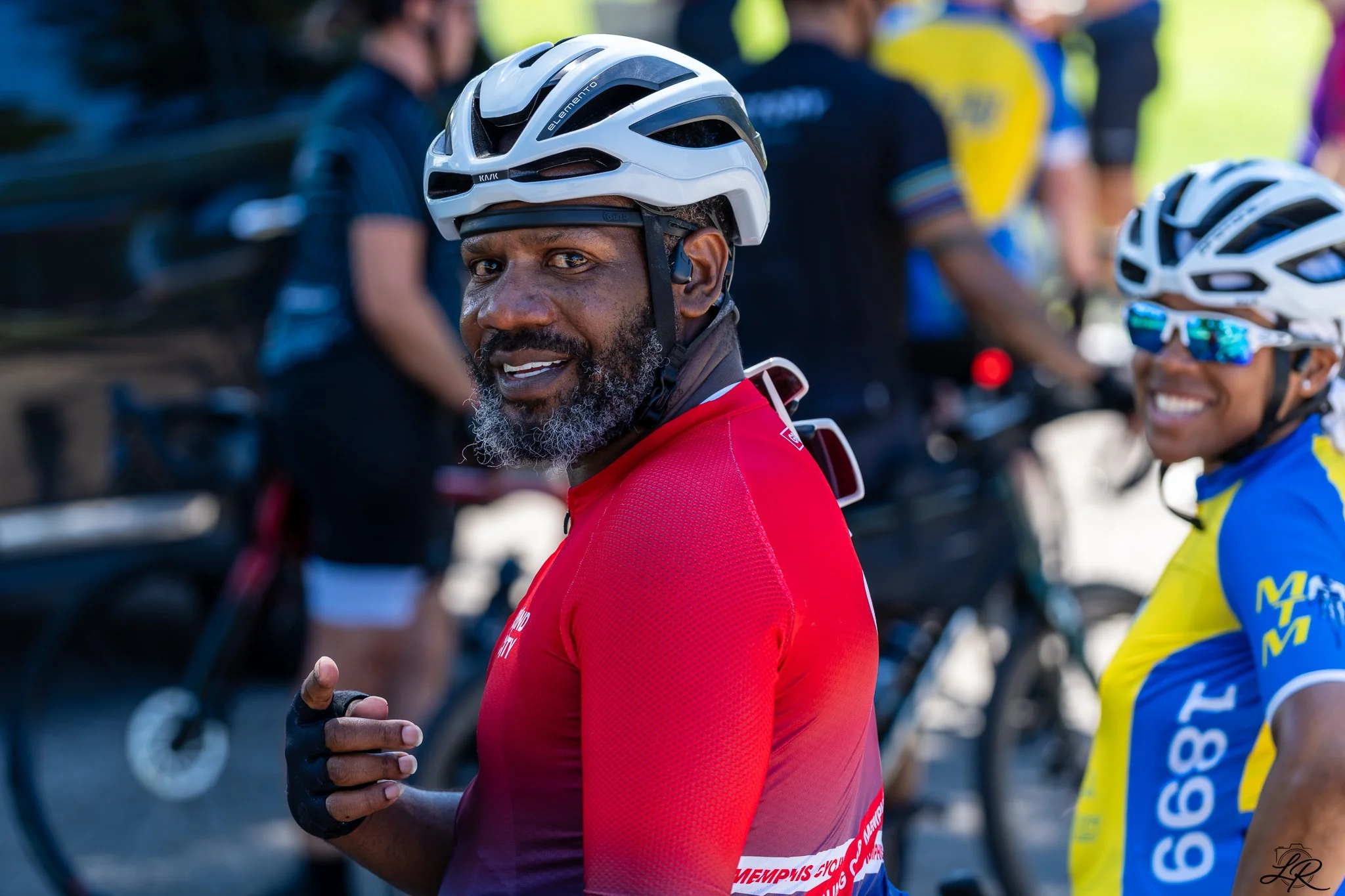 A man with a beard, wearing a white bicycle helmet and a red cycling jersey, is smiling and looking to the side during a cycling event. There are other cyclists in colorful gear and helmets in the background.