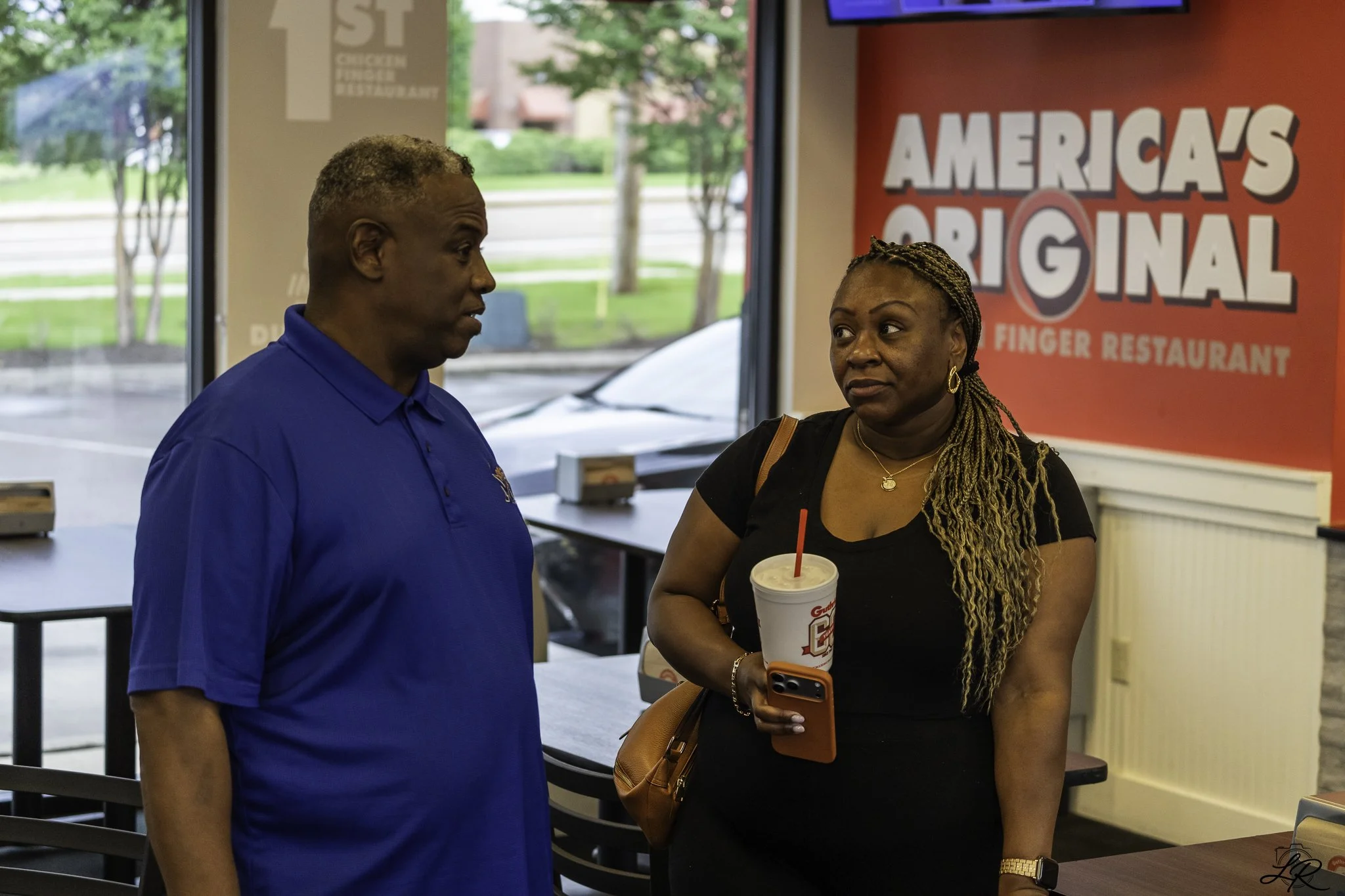 A man in a blue polo shirt and a woman in a black shirt are standing inside a fast-food restaurant, having a conversation. The woman is holding a large drink cup with a red straw, and the man is looking at her. There is a red sign in the background t