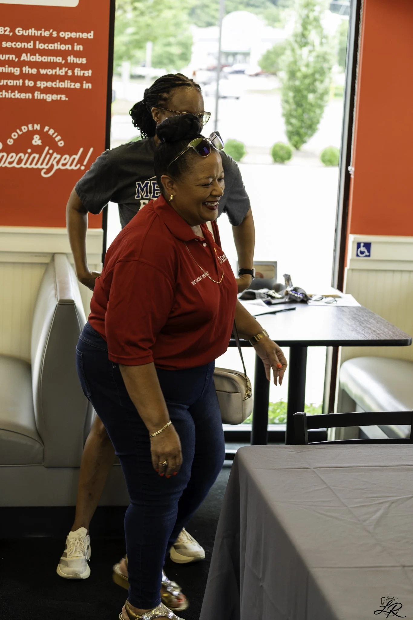 Two women, one wearing a red polo shirt and the other in a dark T-shirt, are laughing and sharing a moment inside a restaurant or cafe with a red and white color scheme. One woman is leaning on the other with a big smile, and there is a table with a 