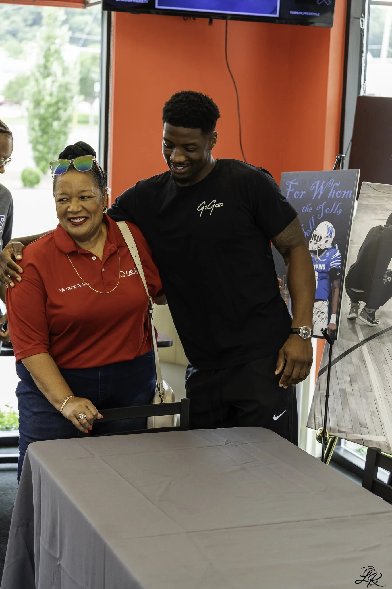 A woman and a young man are smiling and hugging behind a table covered with a grey tablecloth in a room with large windows and red walls. A sign in the background reads "For Whom the Jolls" with a sports helmet illustration. The woman is wearing a re