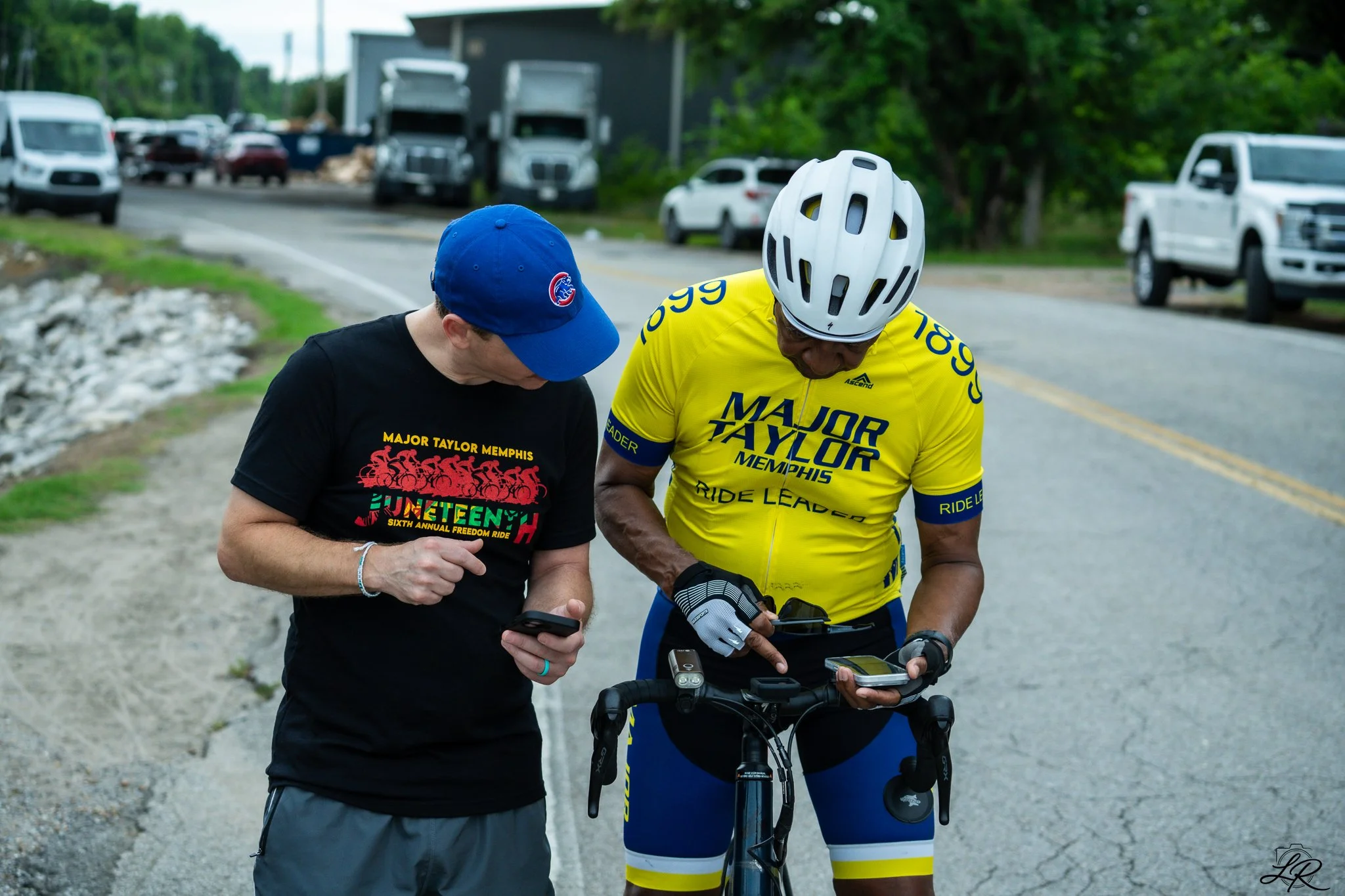 Two men in cycling gear and a helmet look at their phones while standing with their bikes on a rural road, with parked trucks and trees in the background.