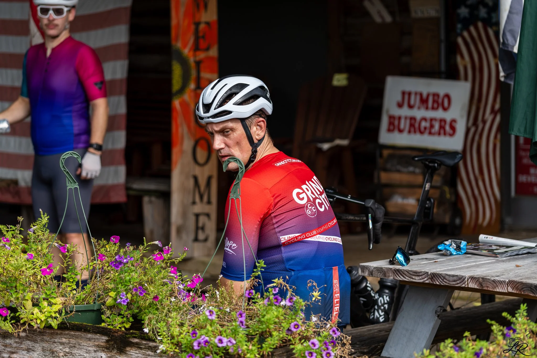 A man wearing a white bike helmet and a red, purple, and blue cycling jersey, sitting near a table with bicycles and tools, surrounded by purple flowers, at a shop or café with a sign that reads 'JUMBO BURGERS' and a partially visible person in a col