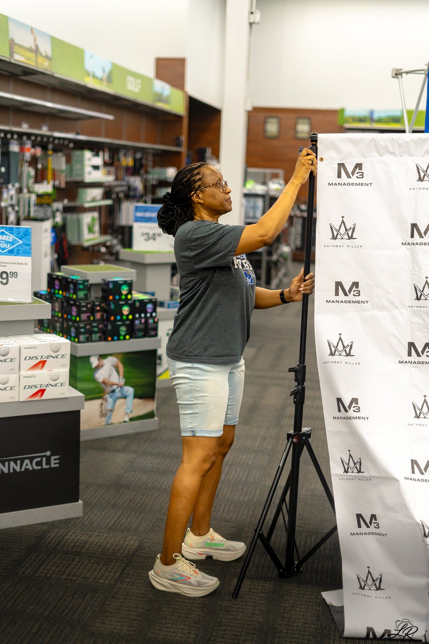 A woman with braided hair and glasses is standing in a store, adjusting a photography backdrop with the logo 'M3 Management' and 'Anthony Miller.' She is wearing a gray T-shirt, light denim shorts, and sneakers with green accents.