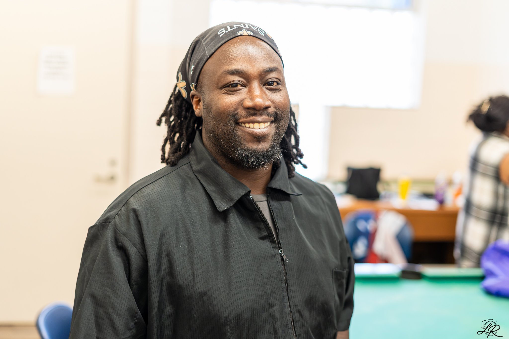 A smiling African American man with dreadlocks, wearing a black jacket and a Saints headscarf, standing in a room with chairs and sports equipment in the background.