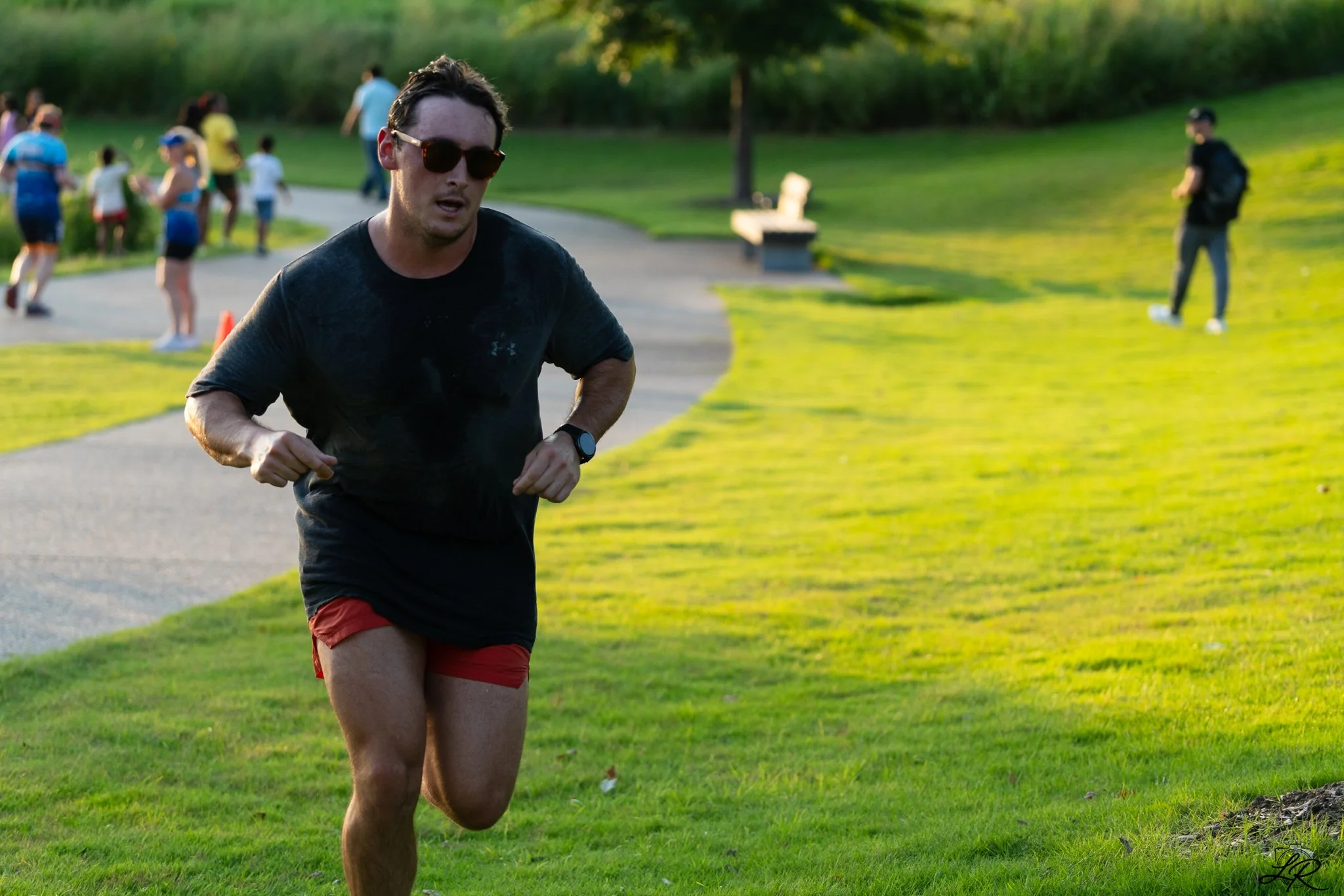 A man running in a park with green grass and trees, wearing sunglasses, a black t-shirt, and red shorts, during daytime with sunlight.