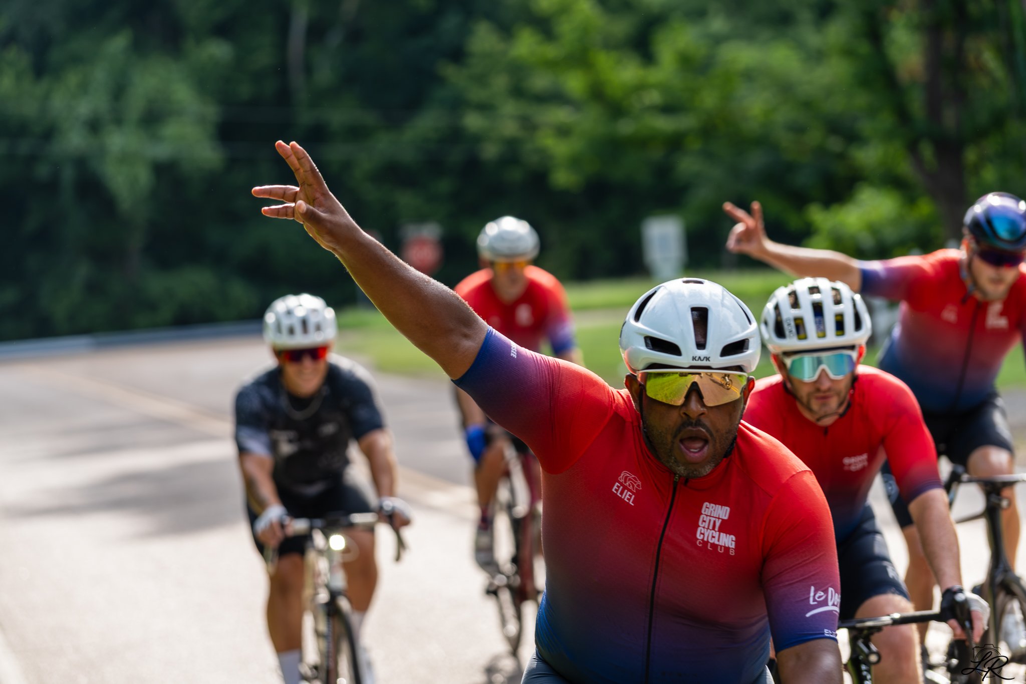 A group of cyclists wearing helmets ride together outdoors on a sunny day, with a wooded area in the background.