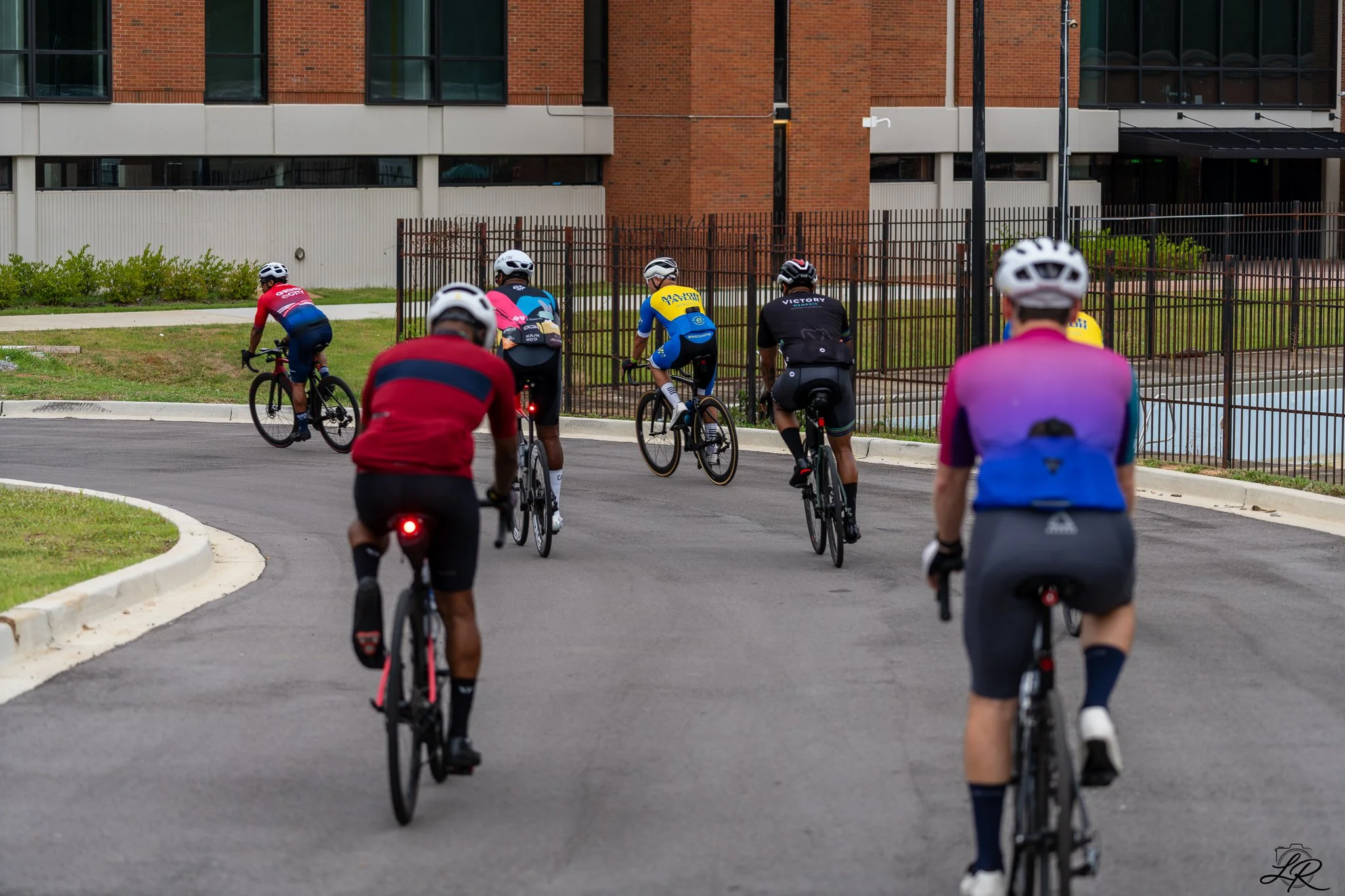 Group of cyclists riding on a curved city road next to a fence and a brick building.