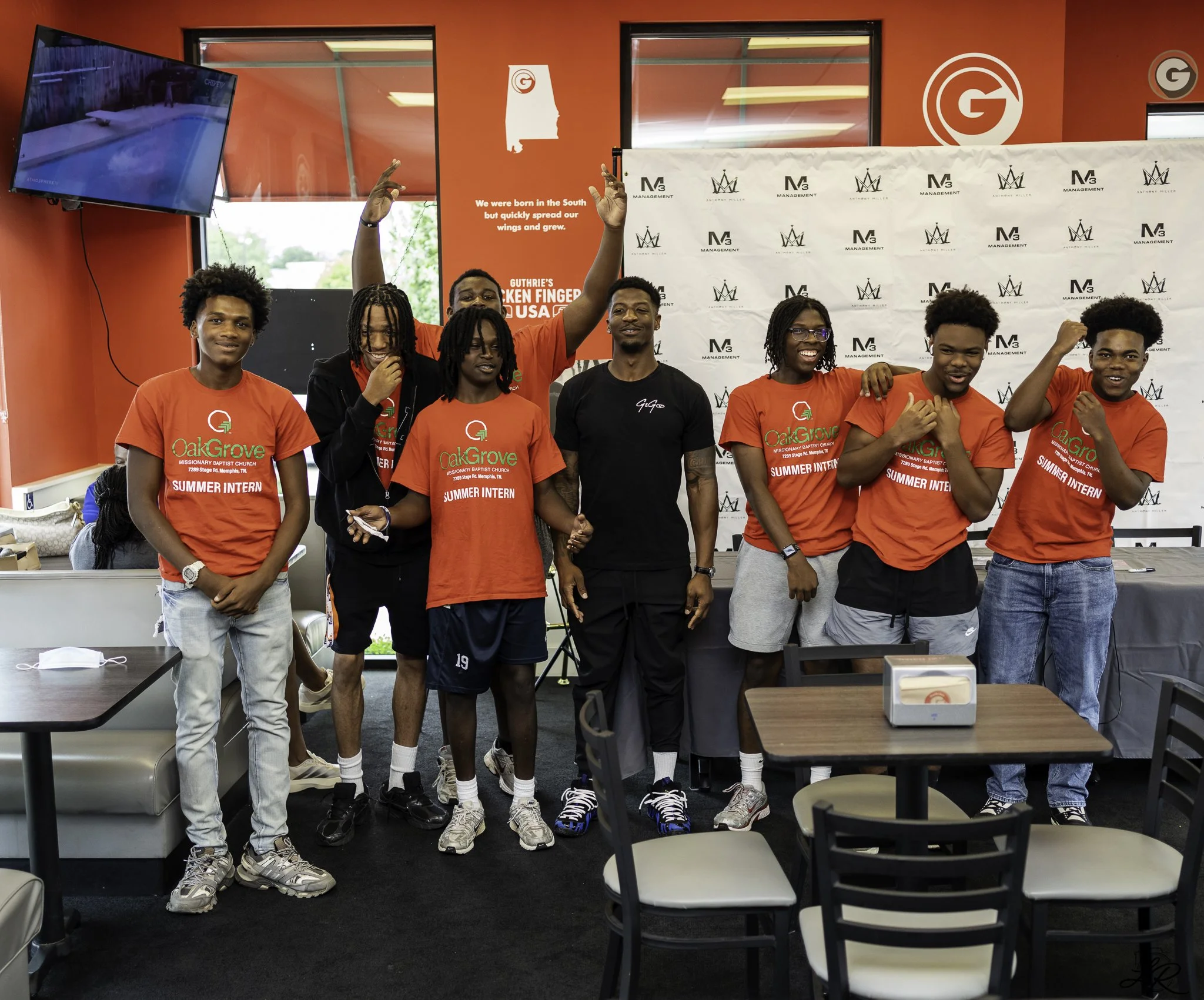 Group of seven young people standing together inside a room with red walls, some wearing red t-shirts that read 'OakGrove Missionary Baptist Church Summer Intern,' with a backdrop displaying logos and a message about South-born wings spreading. They 