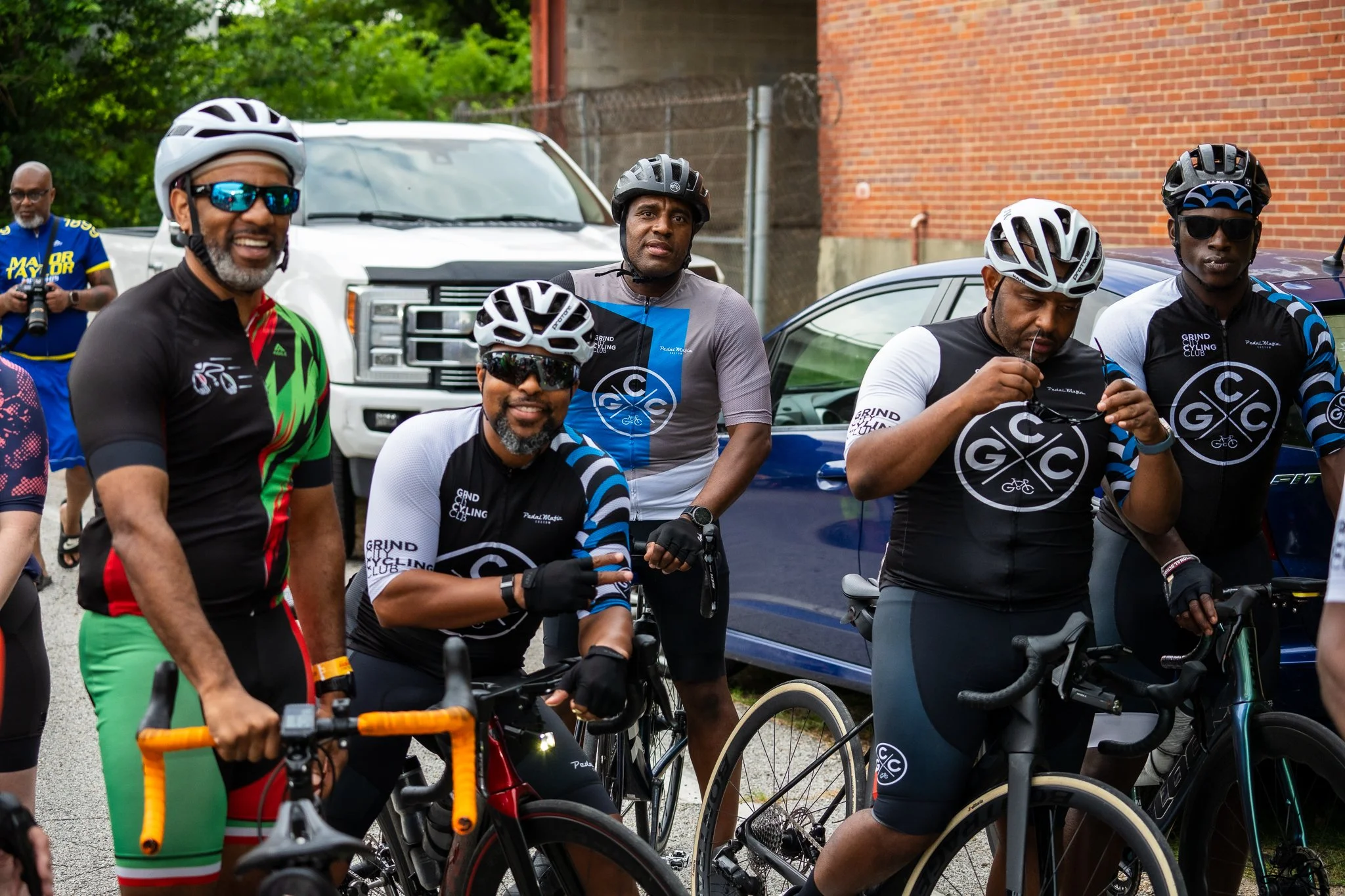Group of diverse cyclists wearing helmets and sunglasses, standing with their bikes outdoors near parked cars and a brick building, some adjusting their gear and others looking toward the camera.