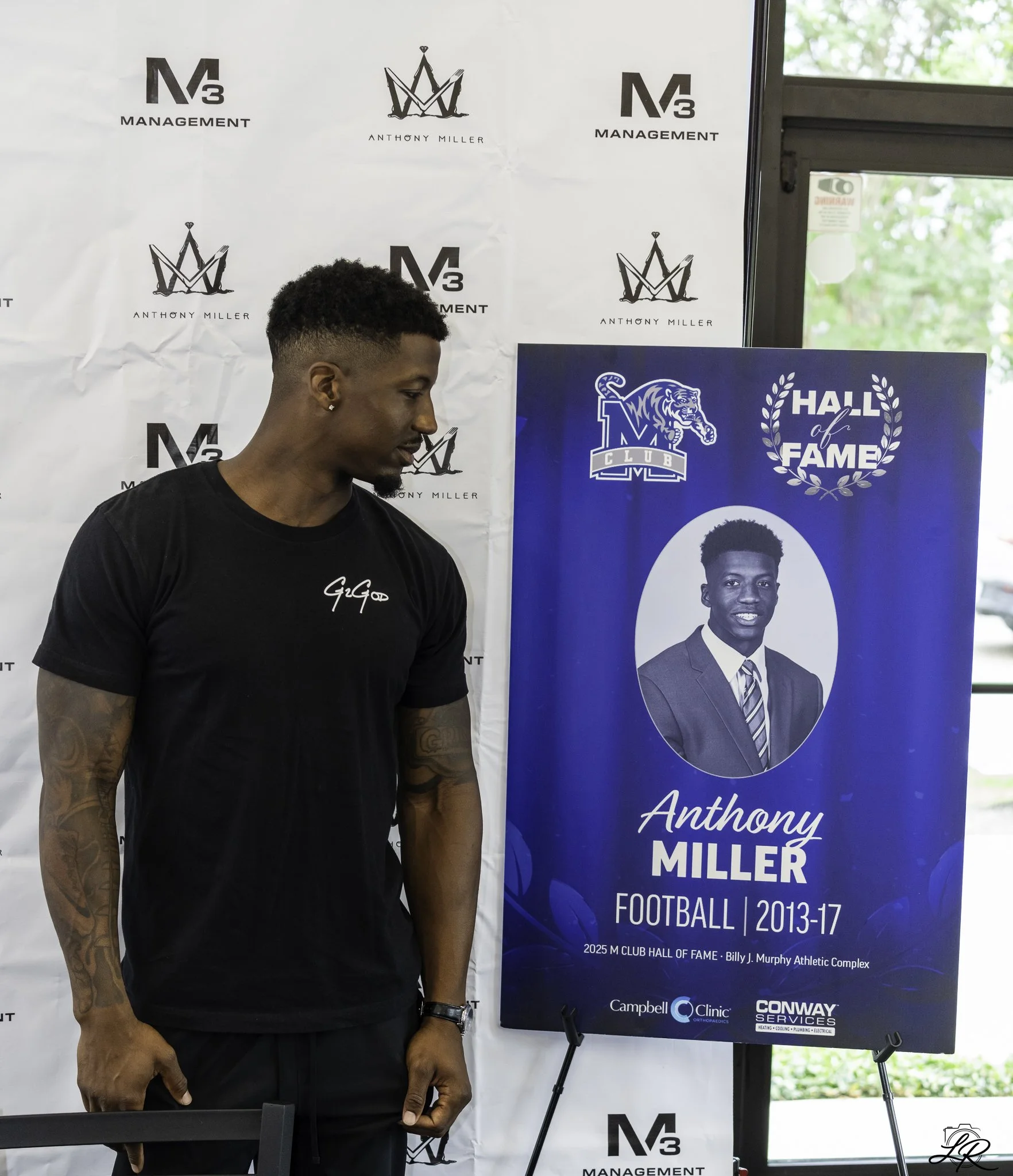 A young man with short curly hair, wearing a black t-shirt, is looking downwards at a table. Behind him is a large blue poster that reads "Hall of Fame" with a photo of a young man in a suit and tie. The background features a white wall with logos of