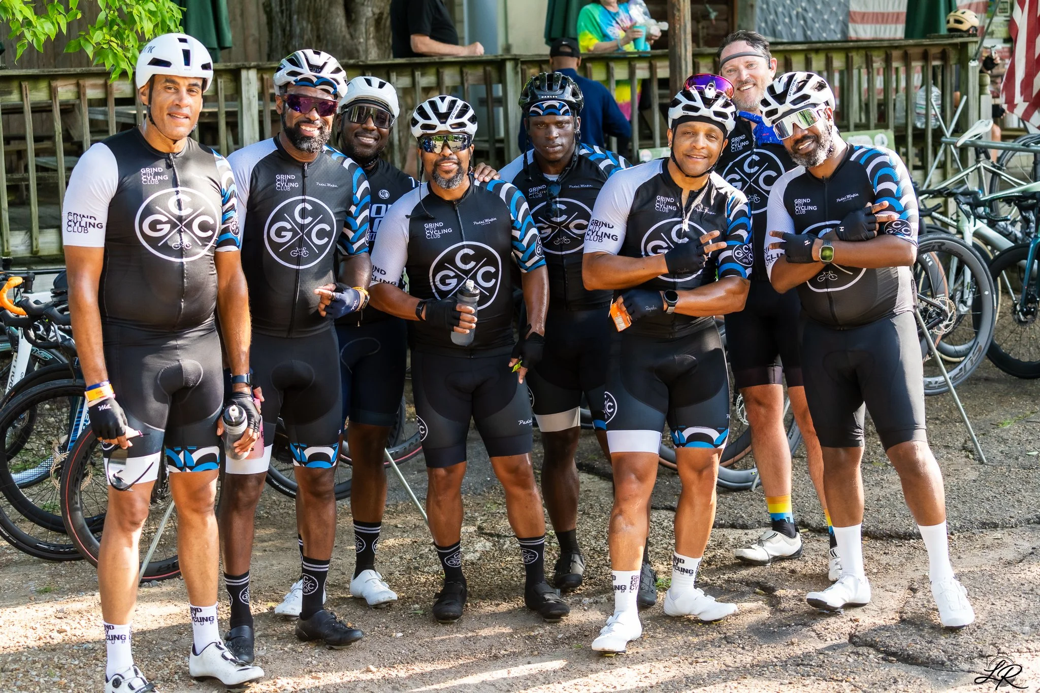 A group of nine men in cycling gear, wearing helmets and sunglasses, posing outdoors with bicycles behind them. They are dressed in black and white cycling jerseys with blue accents and the logo 'GIC' and 'Grinding City Cycling Club.' Some are smilin