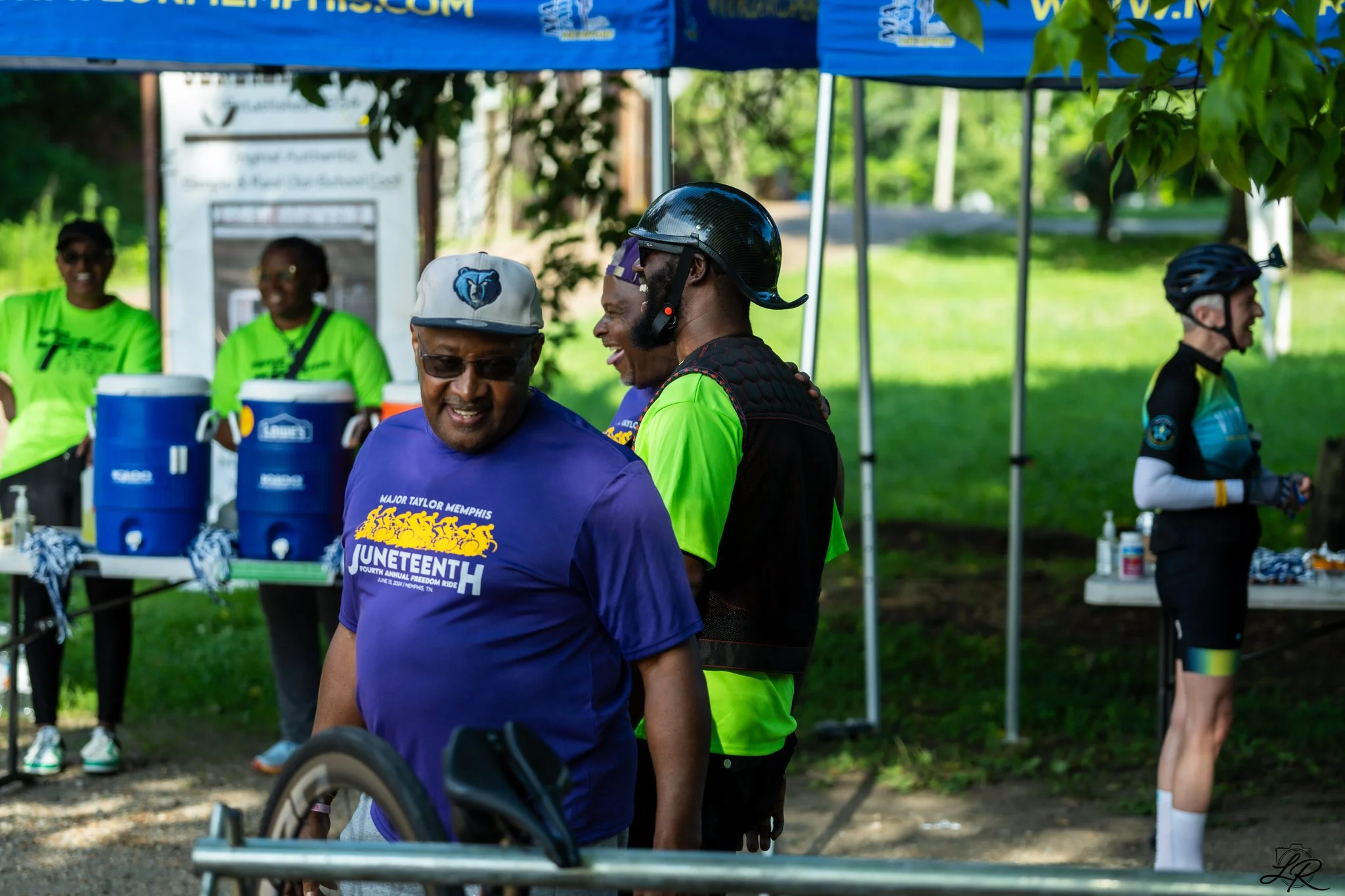 People gathered outdoors at a community event, some wearing bright green shirts with a Memphis theme, under a blue canopy with water coolers on a table. Two men are smiling and talking, one wearing a Memphis hat and the other wearing a helmet, with o