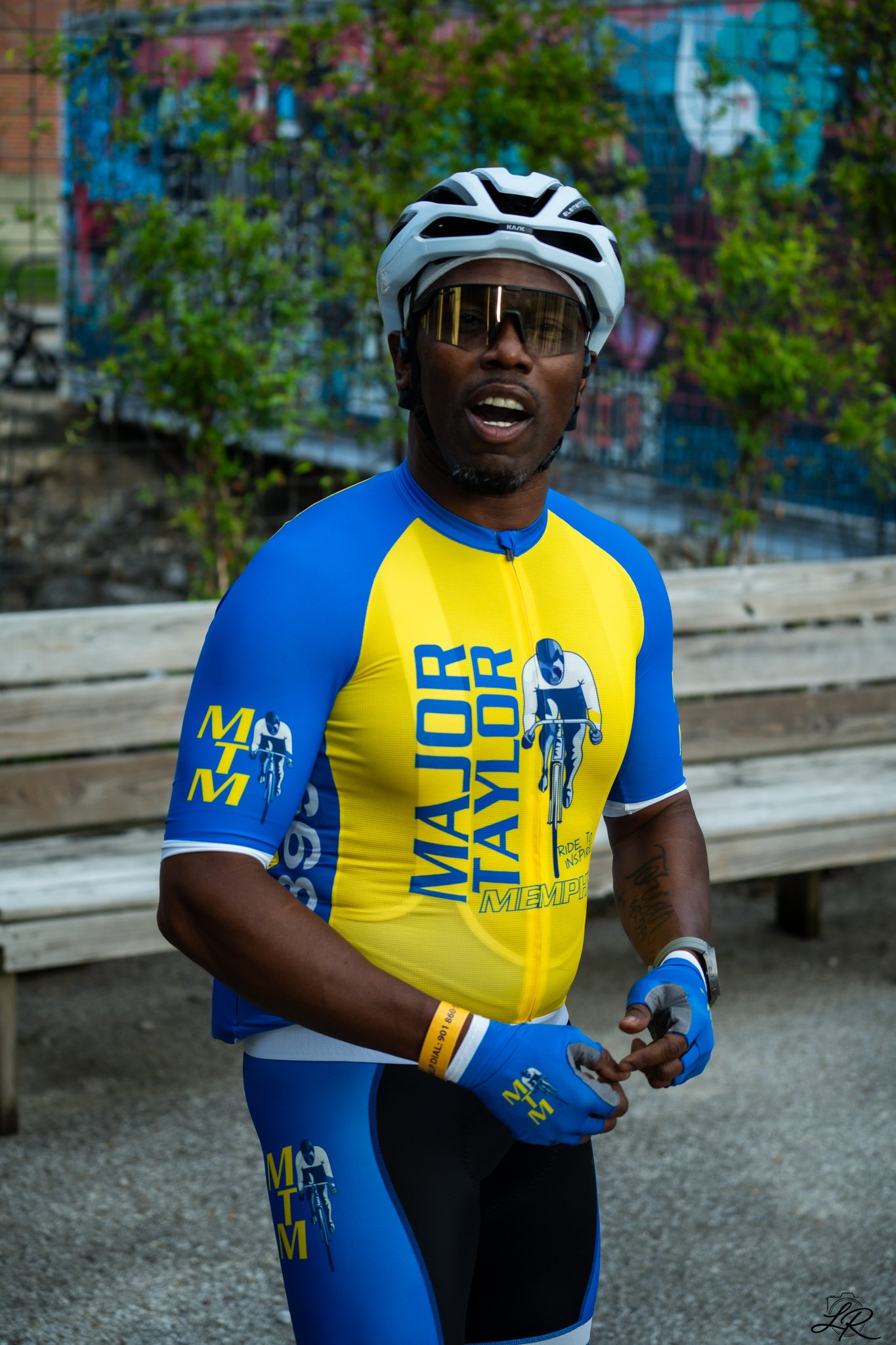 Man wearing a blue and yellow cycling jersey and gloves, white helmet and sunglasses, standing outdoors with trees and a building in the background.
