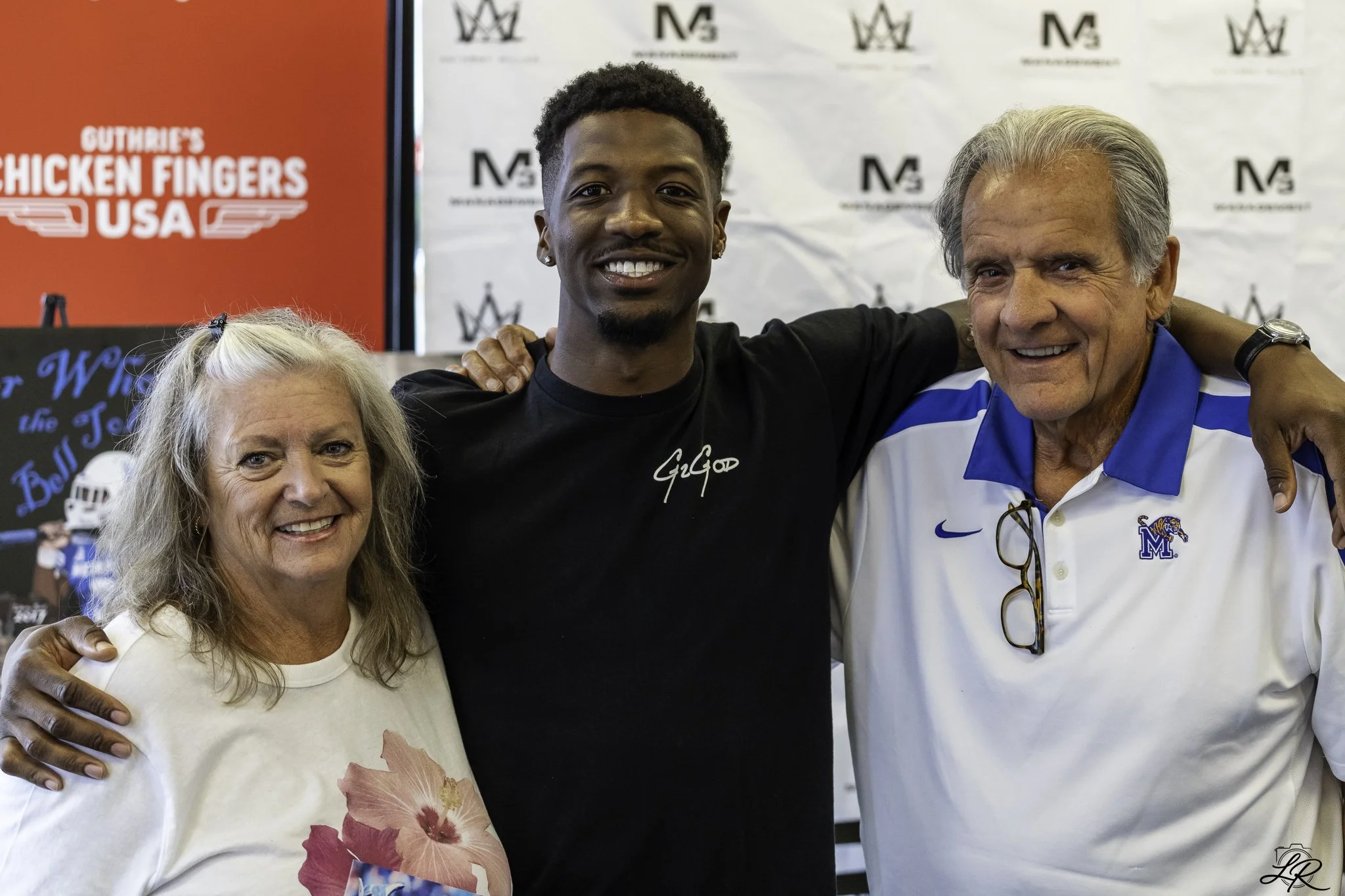A group photo of three people smiling, with a young man in the center and an older woman and man on either side, taken at an indoor event with a backdrop featuring logos and a red sign in the background.