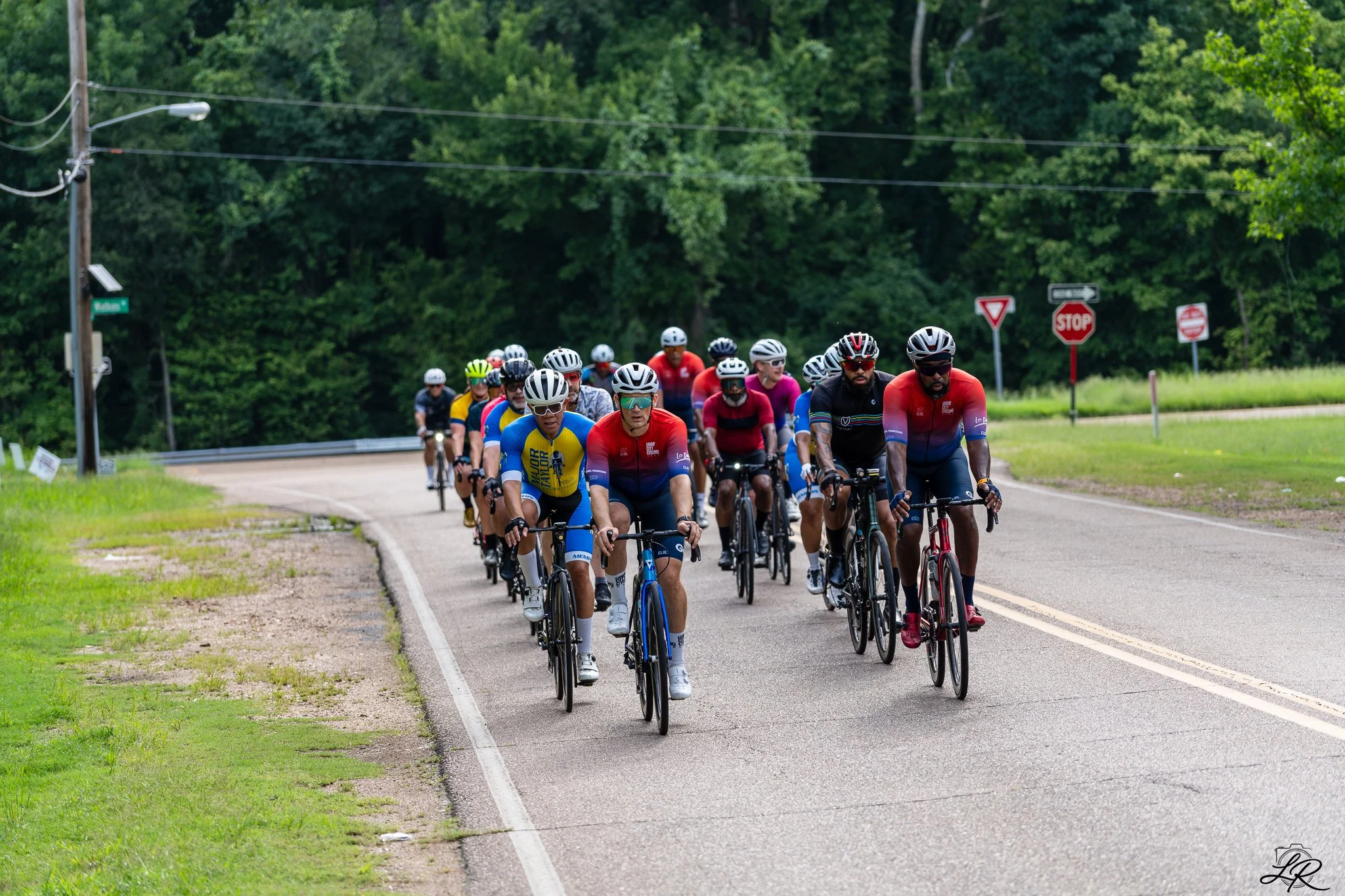 A large group of cyclists riding together on a rural road surrounded by green trees and grass.