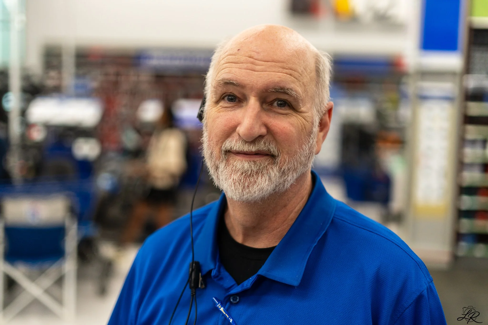 Portrait of an older man with a white beard and bald head wearing a blue shirt, standing in what appears to be a retail store or office with shelves in the background.