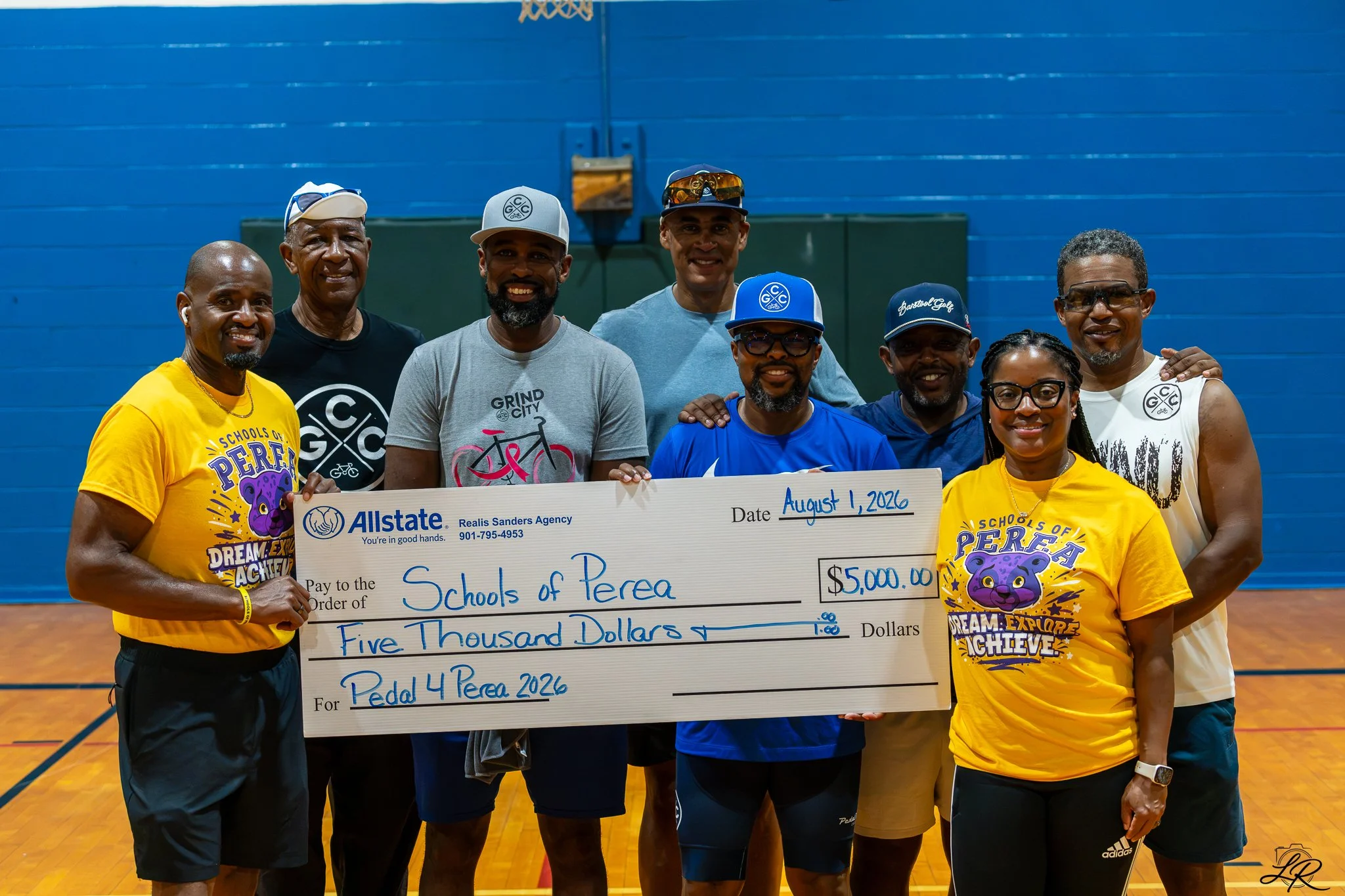 Group of eight diverse adults standing inside a gymnasium, holding a large check made out to 'Schools of Perea' for five thousand dollars, dated August 1, 2026, with some smiling and wearing casual sportswear and sunglasses.