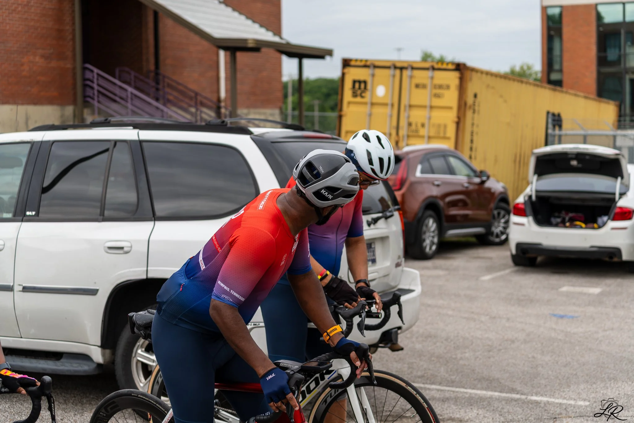 Two cyclists wearing helmets and racing jerseys crouched over their bikes in a parking lot, with multiple parked cars and a yellow shipping container in the background.