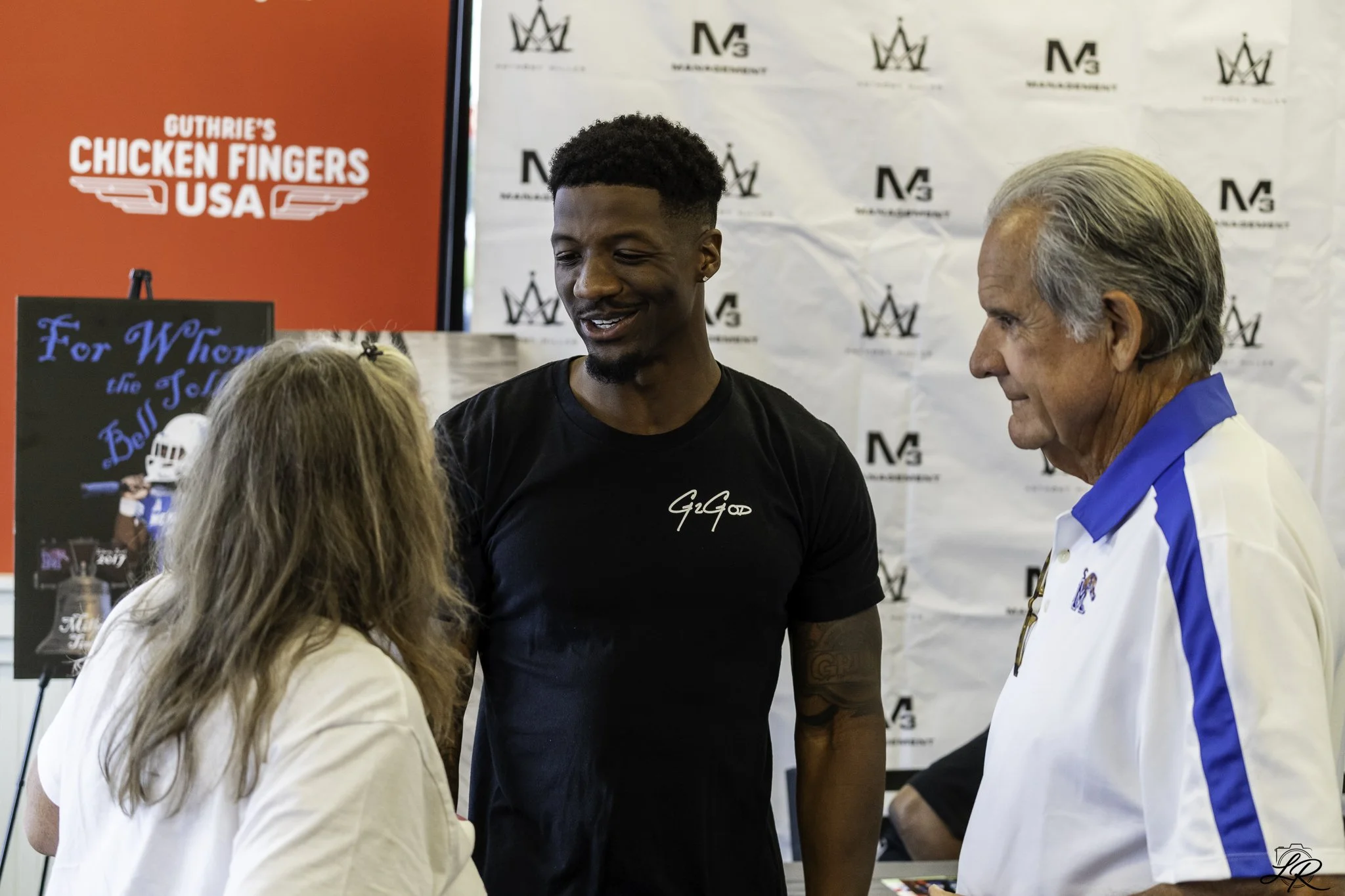 Three people are engaged in conversation at an event. The man in the center has dark skin, short curly hair, and is wearing a black shirt with white text. An older man with light skin, gray hair, and a blue-and-white sports jacket is on the right. A 