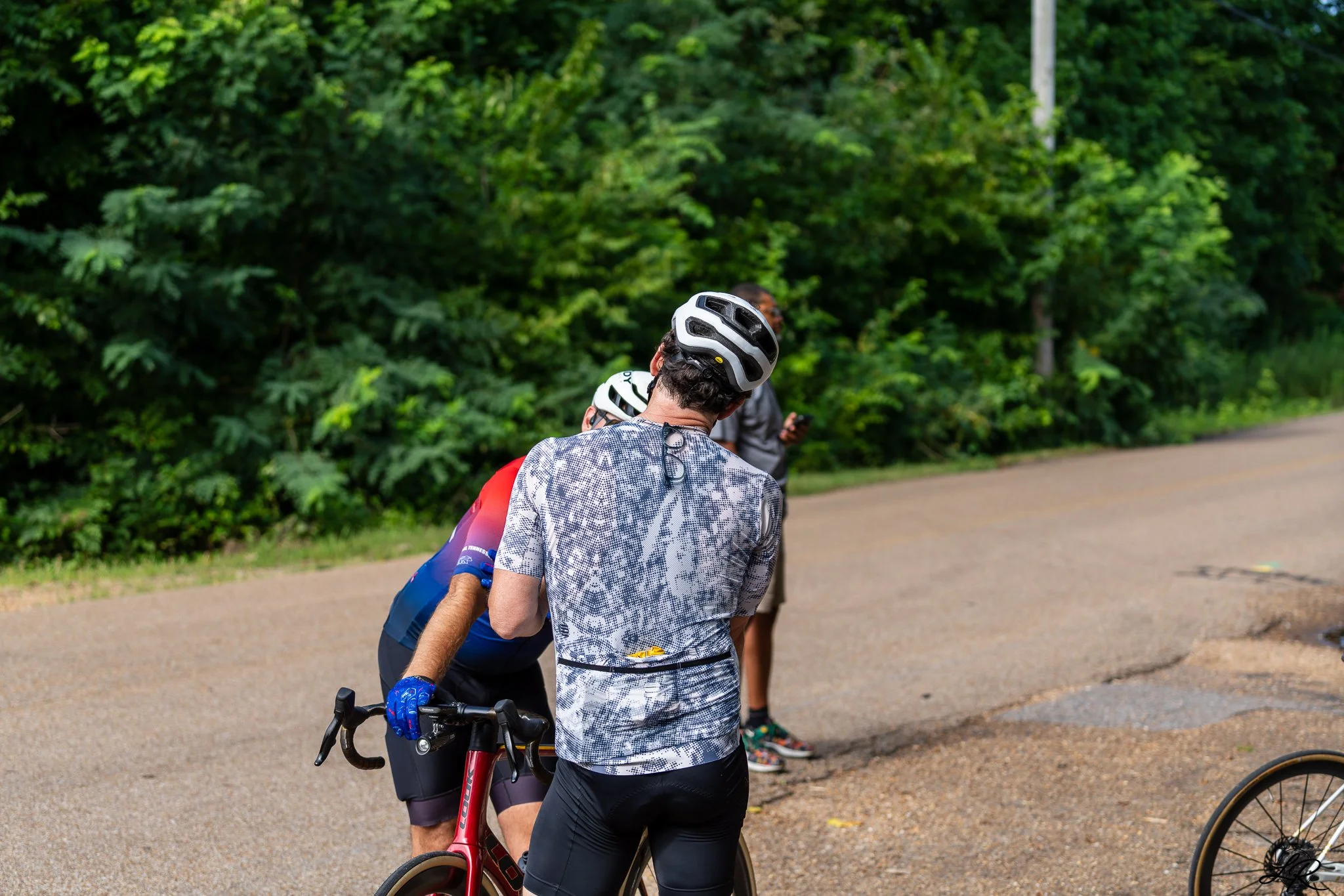 Multiple cyclists wearing helmets and athletic clothing gathered outdoors on a paved path surrounded by green trees.