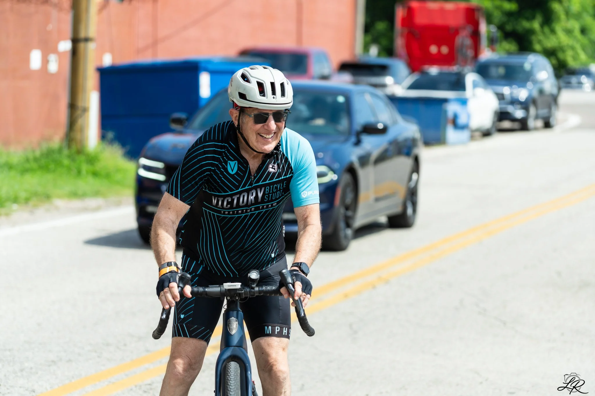 An elderly man riding a bicycle on a street, wearing a white helmet, black sunglasses, and a black and blue cycling jersey, smiling as he pedals.