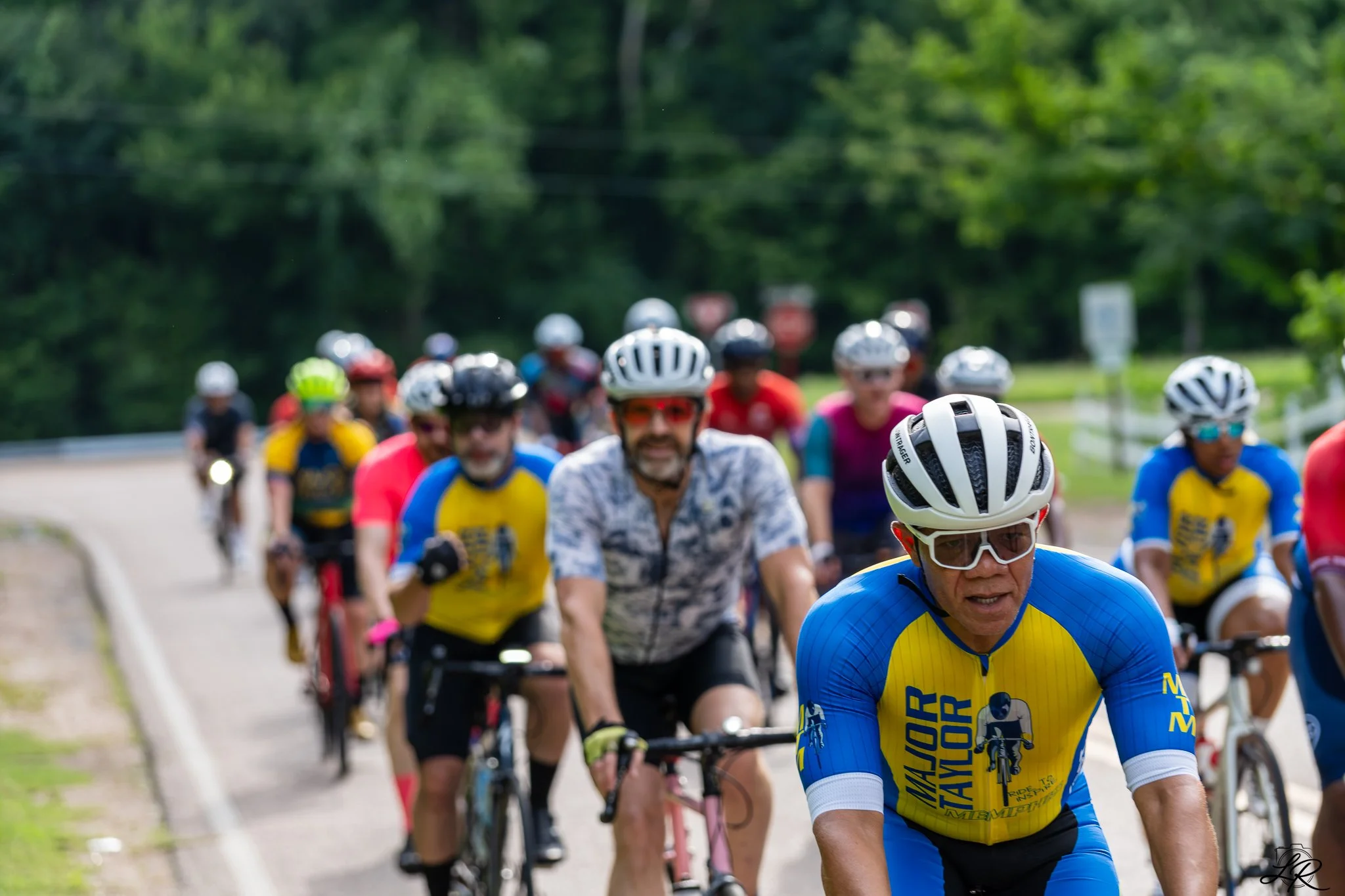 A group of people riding bicycles on a road surrounded by greenery, wearing helmets and colorful sportswear.