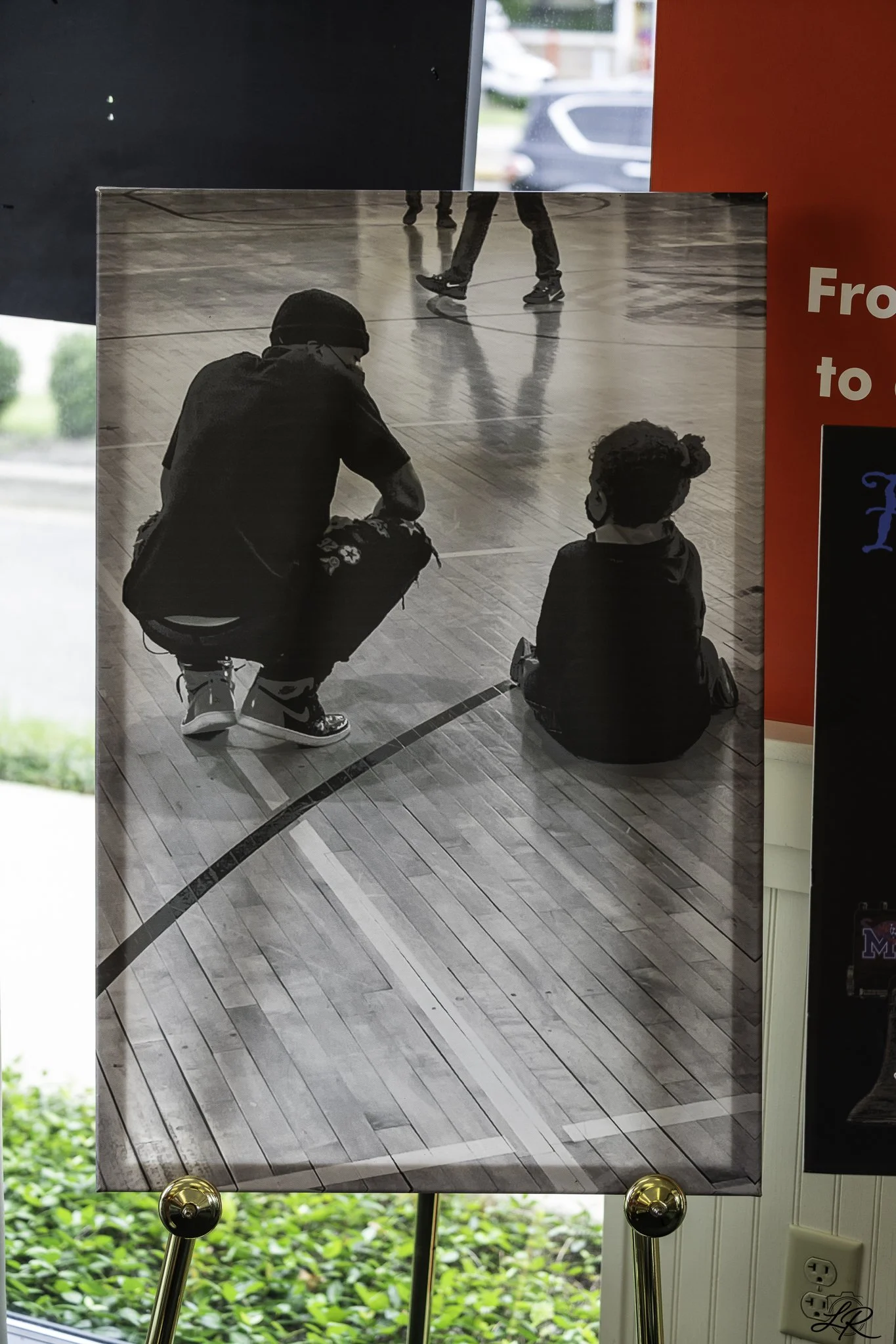 Black and white photo of a person squatting down and talking to a child who is sitting on the floor in a gymnasium or indoor sports facility with a polished wooden floor. The person is wearing a beanie, glasses, a black shirt, and sneakers, while the