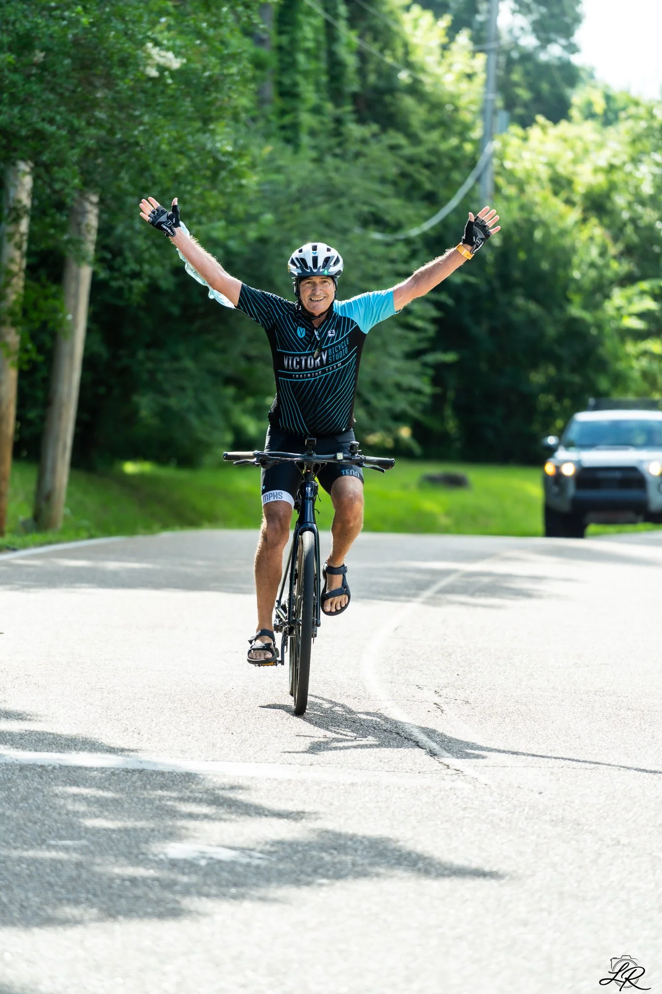 A man riding a bicycle on a sunny road with his arms raised in victory, wearing a helmet and cycling gear, with trees and a car in the background.
