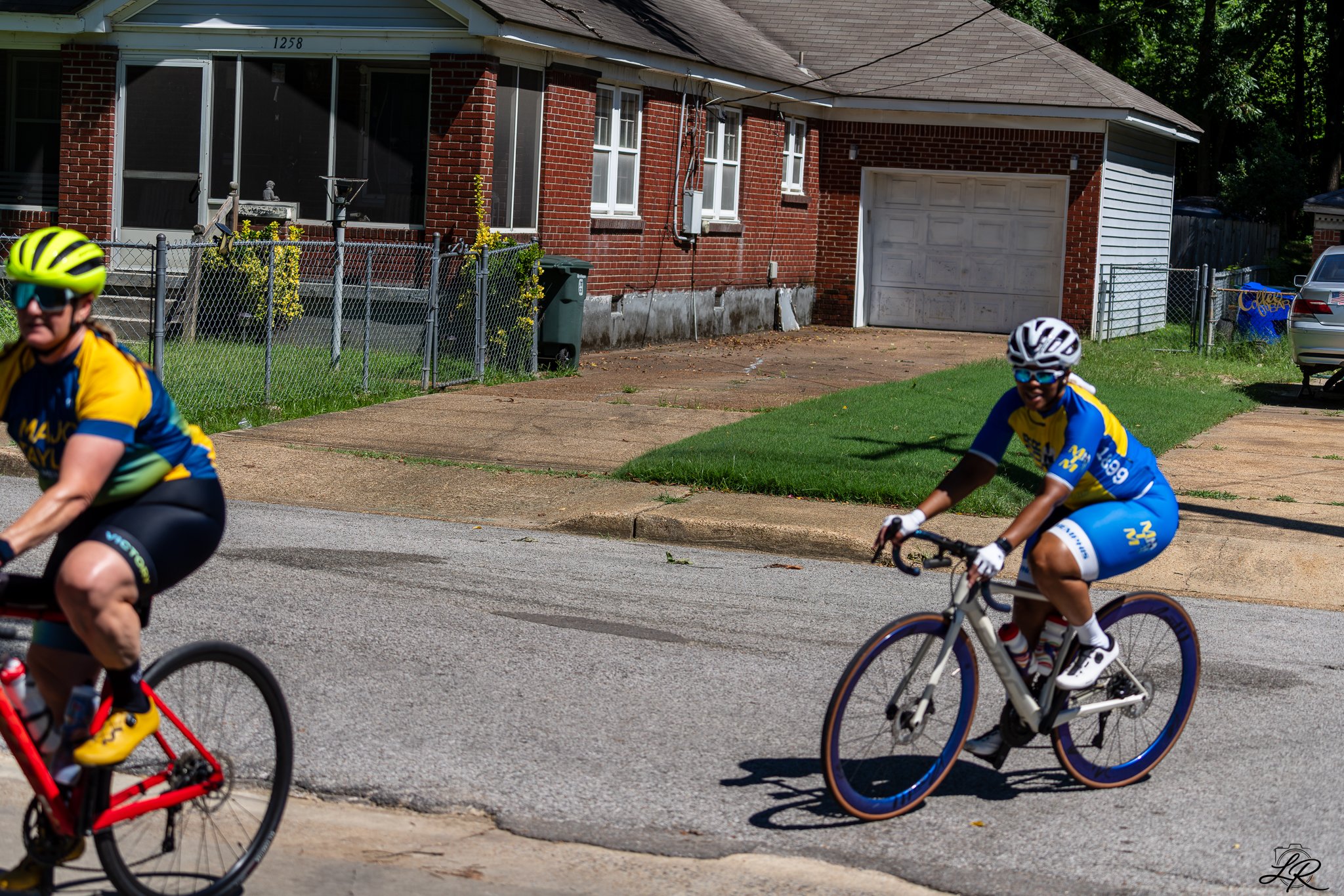Two cyclists wearing blue and yellow uniforms ride their bikes on the street in front of a brick house with a white garage door. The house has a screened porch and a chain-link fence with some plants. The image is taken on a sunny day.