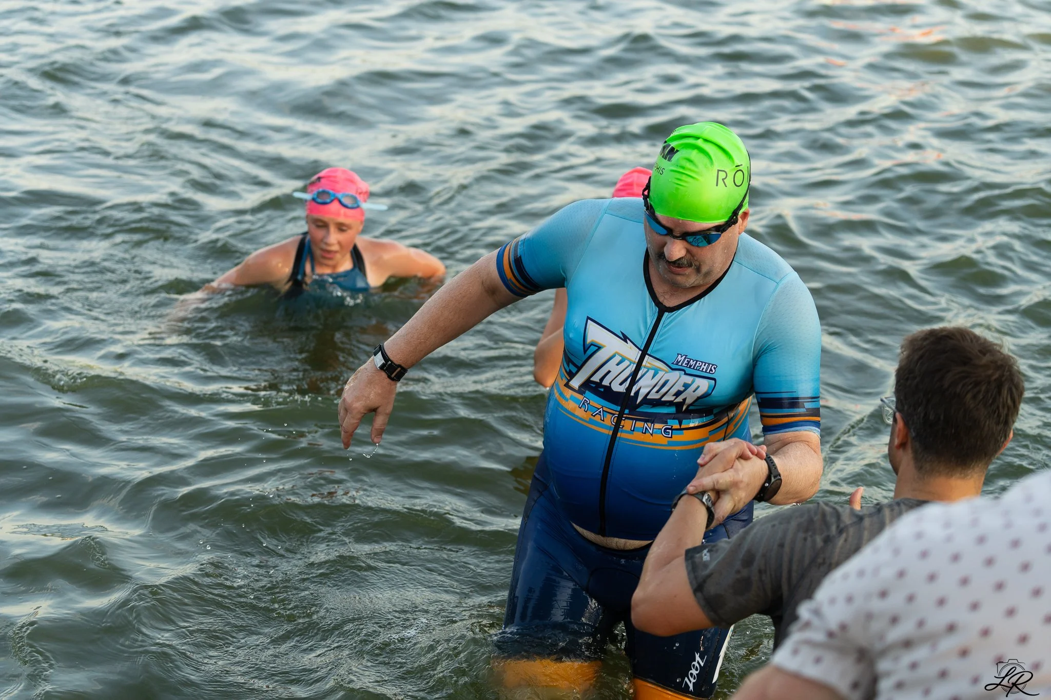 A man in a blue and orange triathlon suit with a green swim cap and sunglasses, helping another person out of the water, while a woman with a pink swim cap and goggles looks on.