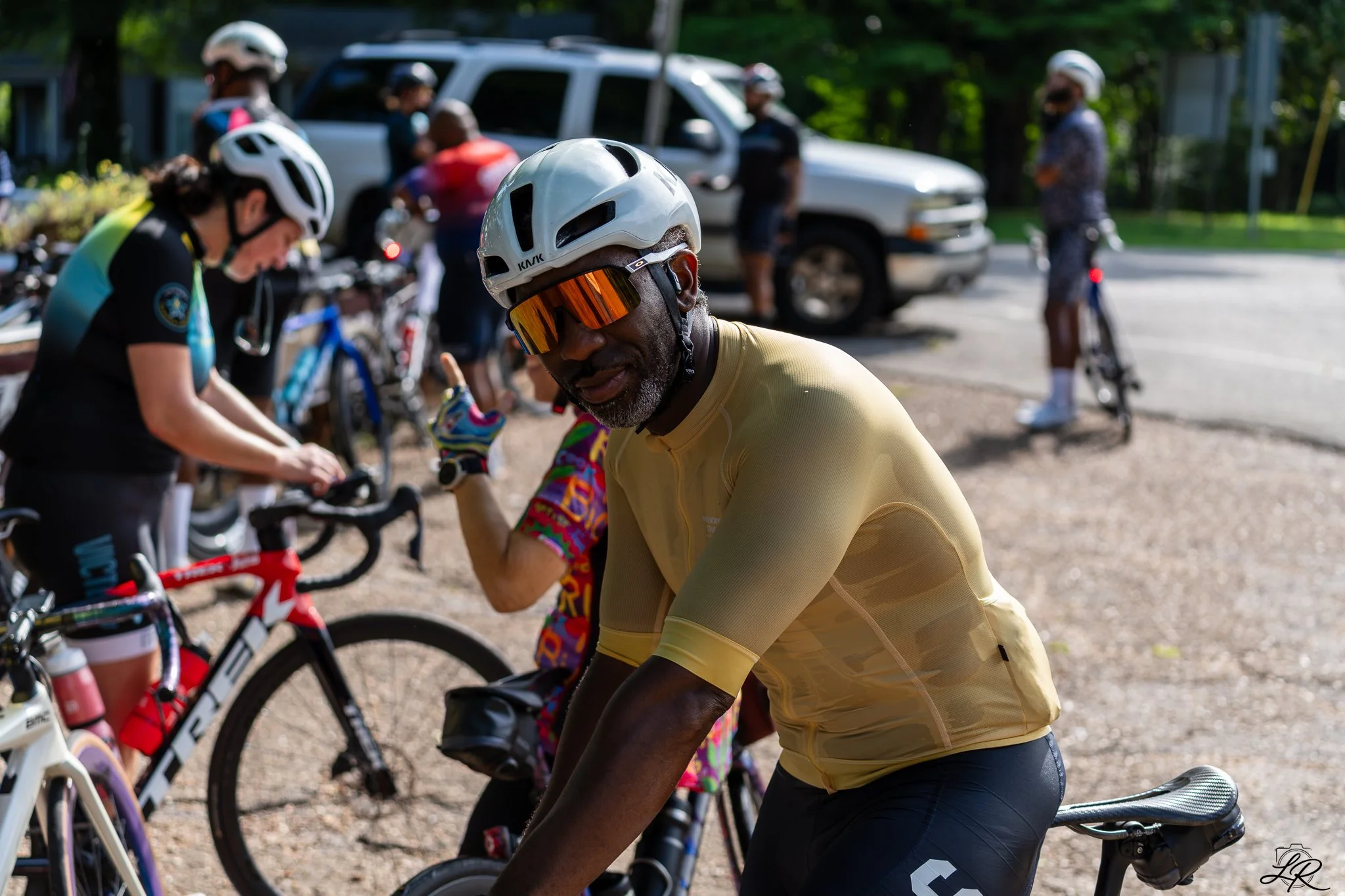 A group of cyclists gathered outdoors, wearing helmets and sports gear, with some people standing next to their bikes and others preparing to ride.