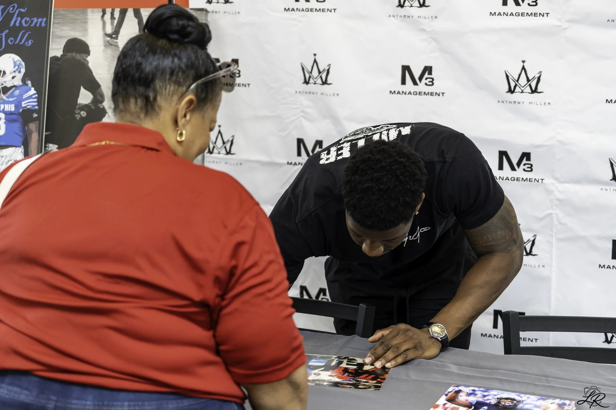 A woman in a red top is standing in front of a table with photos on it, watching an autograph signing. An athlete in black apparel with athletic tattoos is signing an item for her. They are indoors, with a backdrop featuring logos for 'M3 Management'