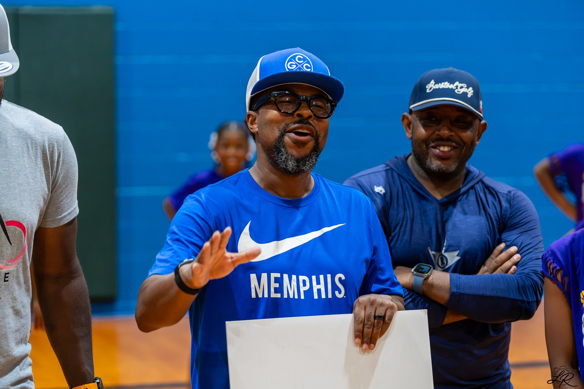 Two men and a woman smiling and talking at an indoor sports event. The man in the center wears glasses, a Memphis shirt, and a blue cap, holding a white sign.