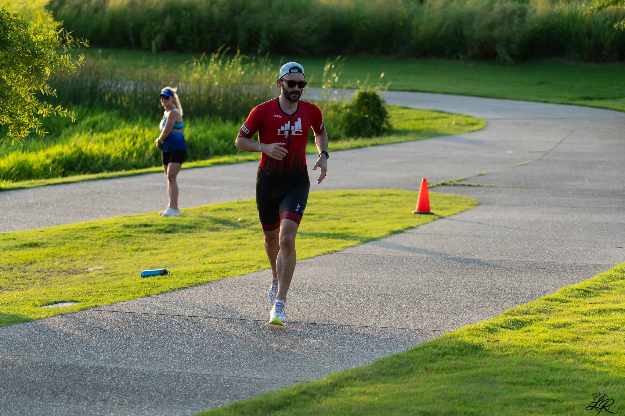 A man in a red athletic shirt, black shorts, white running shoes, and sunglasses running on a curved paved path in a park during late afternoon or early evening. A woman in a blue tank top, black shorts, and sunglasses stands on the grass in the back