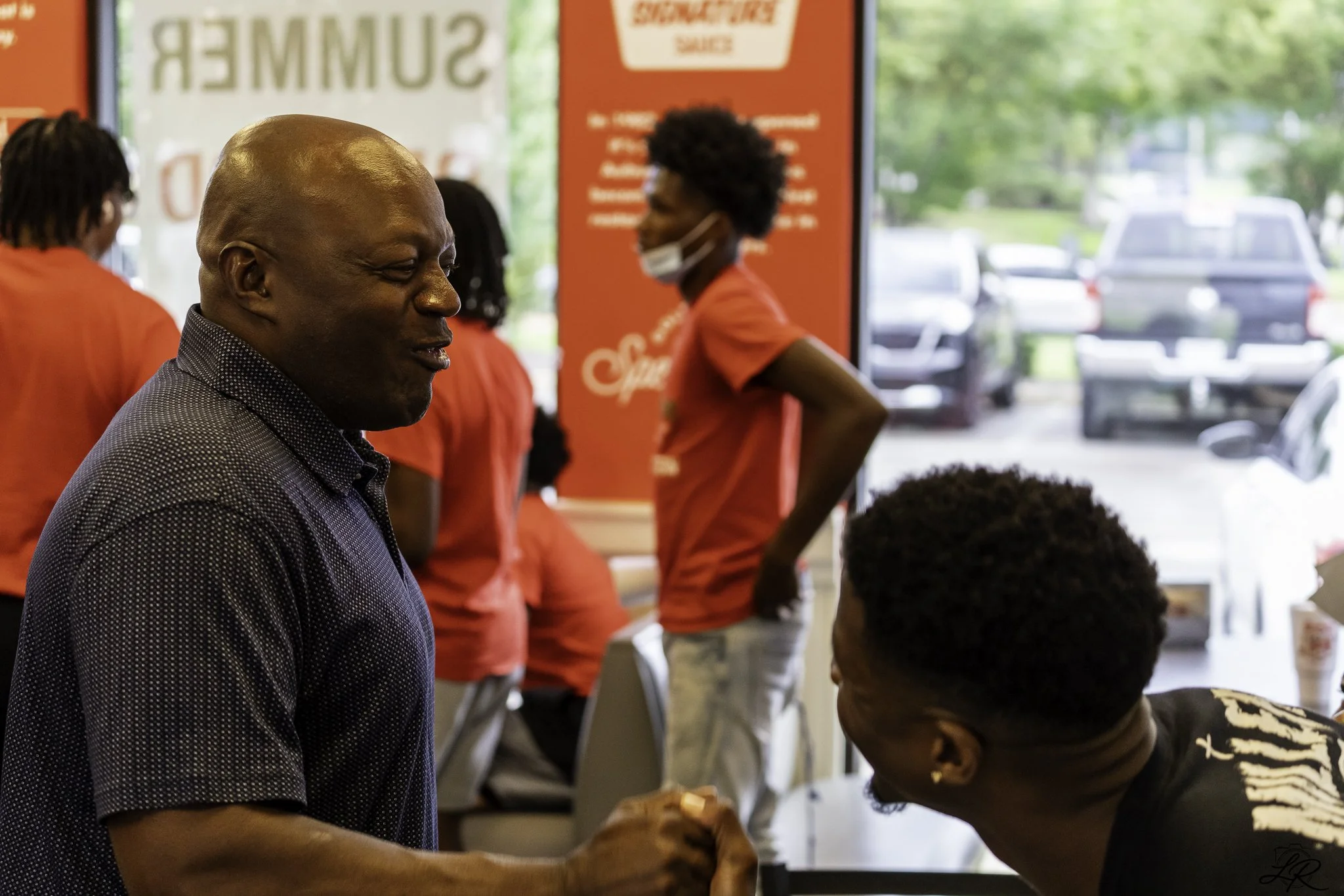 A man with a bald head and a patterned shirt shares a handshake with a young person with short hair, wearing a striped shirt, inside a fast food restaurant. In the background, restaurant staff in red uniforms are visible, with some wearing face masks