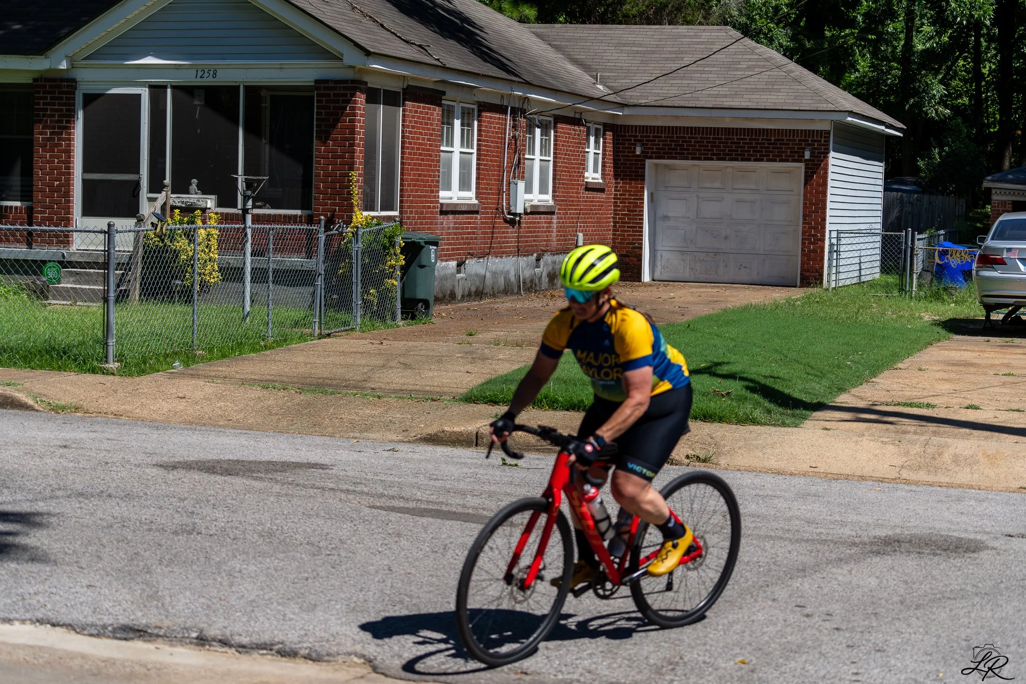 A person riding a red bicycle on a suburban street in front of a brick house with a garage. The rider is wearing a yellow and blue cycling outfit and a yellow helmet.