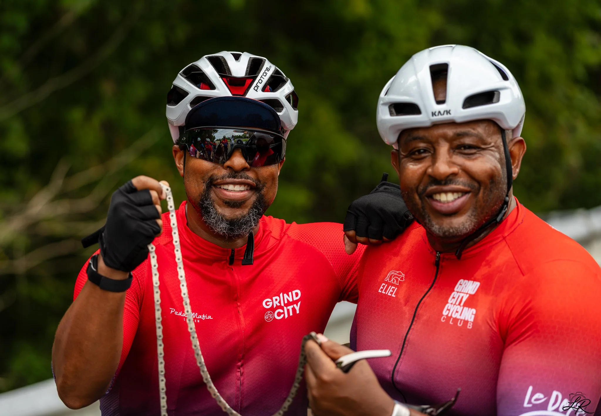 Two smiling men wearing red cycling jerseys and white helmets, standing outdoors with greenery, one pointing at his watch.