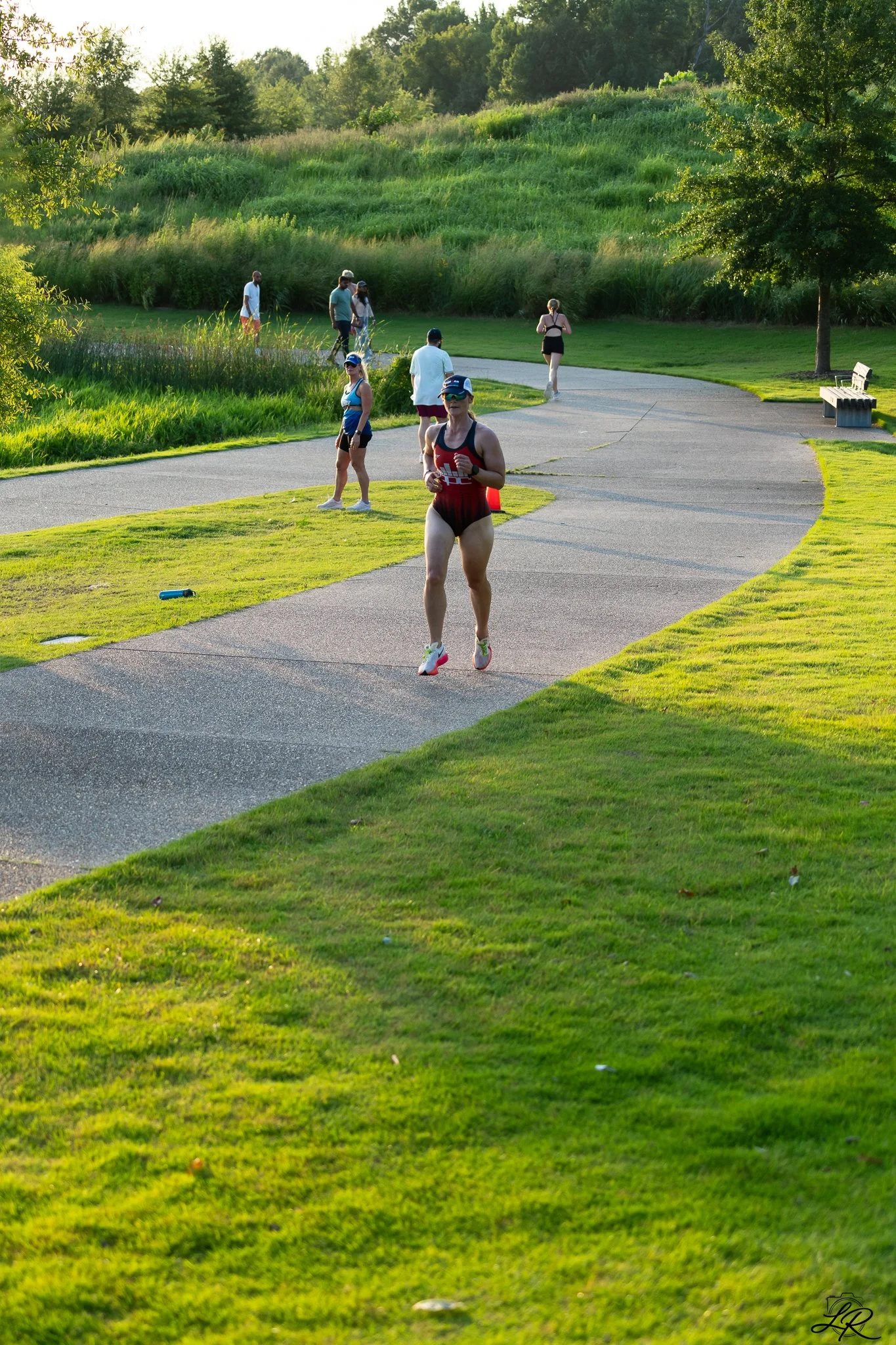 Several people walking and running in a park with green grass, trees, and a curving paved path during daylight.