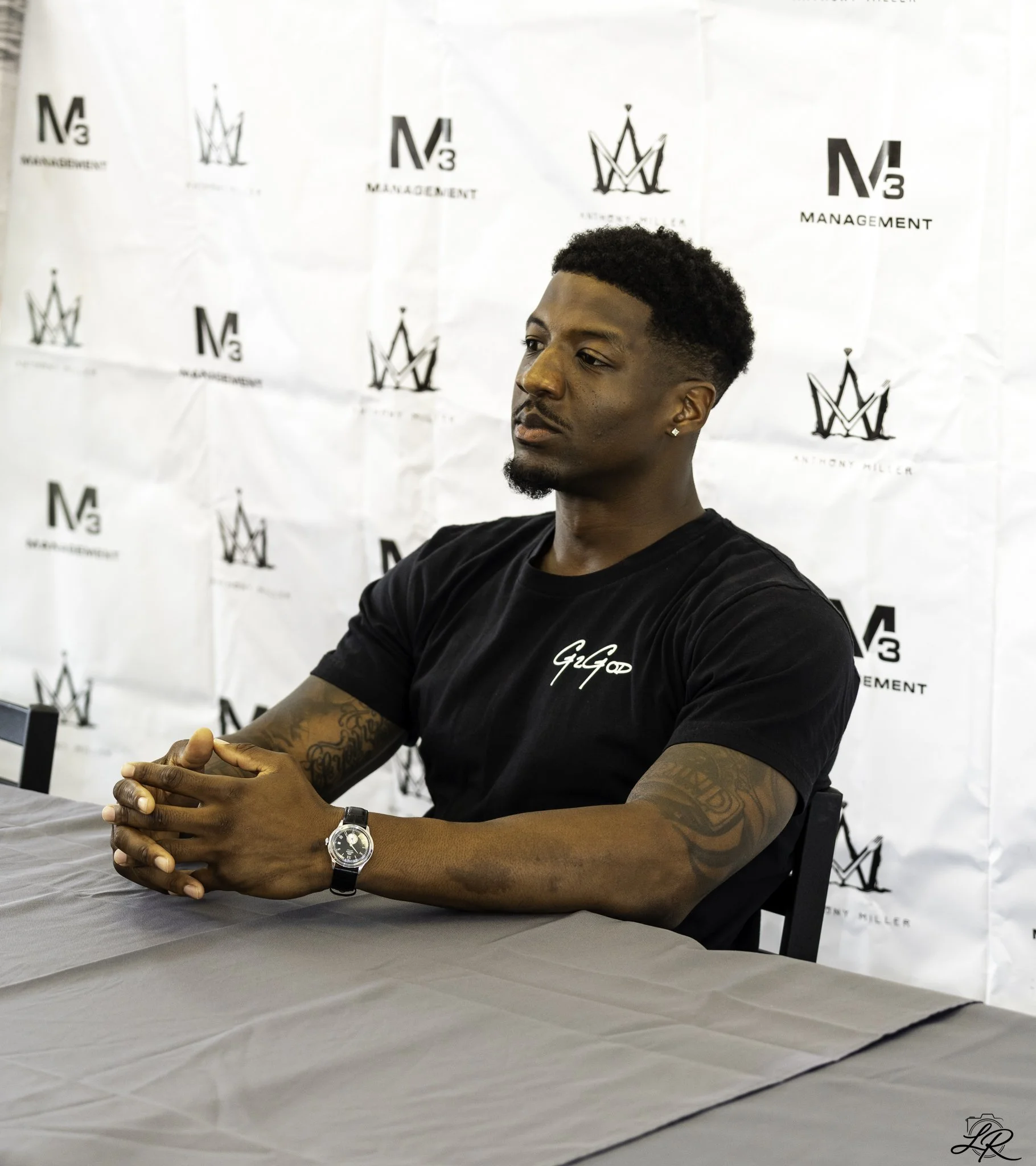 Young man with tattoos on his arm, wearing a black T-shirt and a silver wristwatch, sitting at a table with a gray tablecloth, in front of a white backdrop with logos and text that read 'N3 MANAGEMENT' and 'Anthony Miller.' The man has short curly ha
