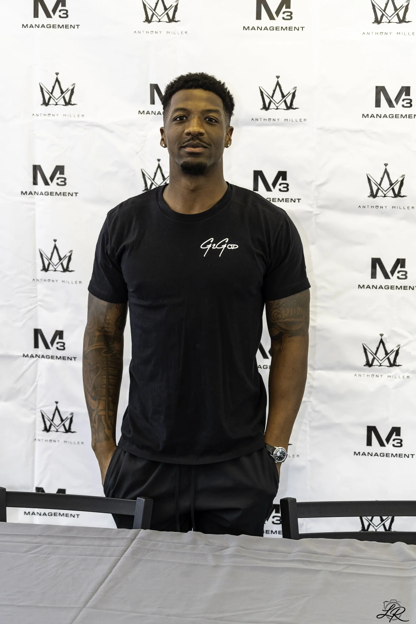 Young man with short curly hair, wearing earrings, a black t-shirt with white text, and a watch on his left wrist, standing in front of a white backdrop with black logos and text for 'N3 Management' and 'Anthony Miller'.