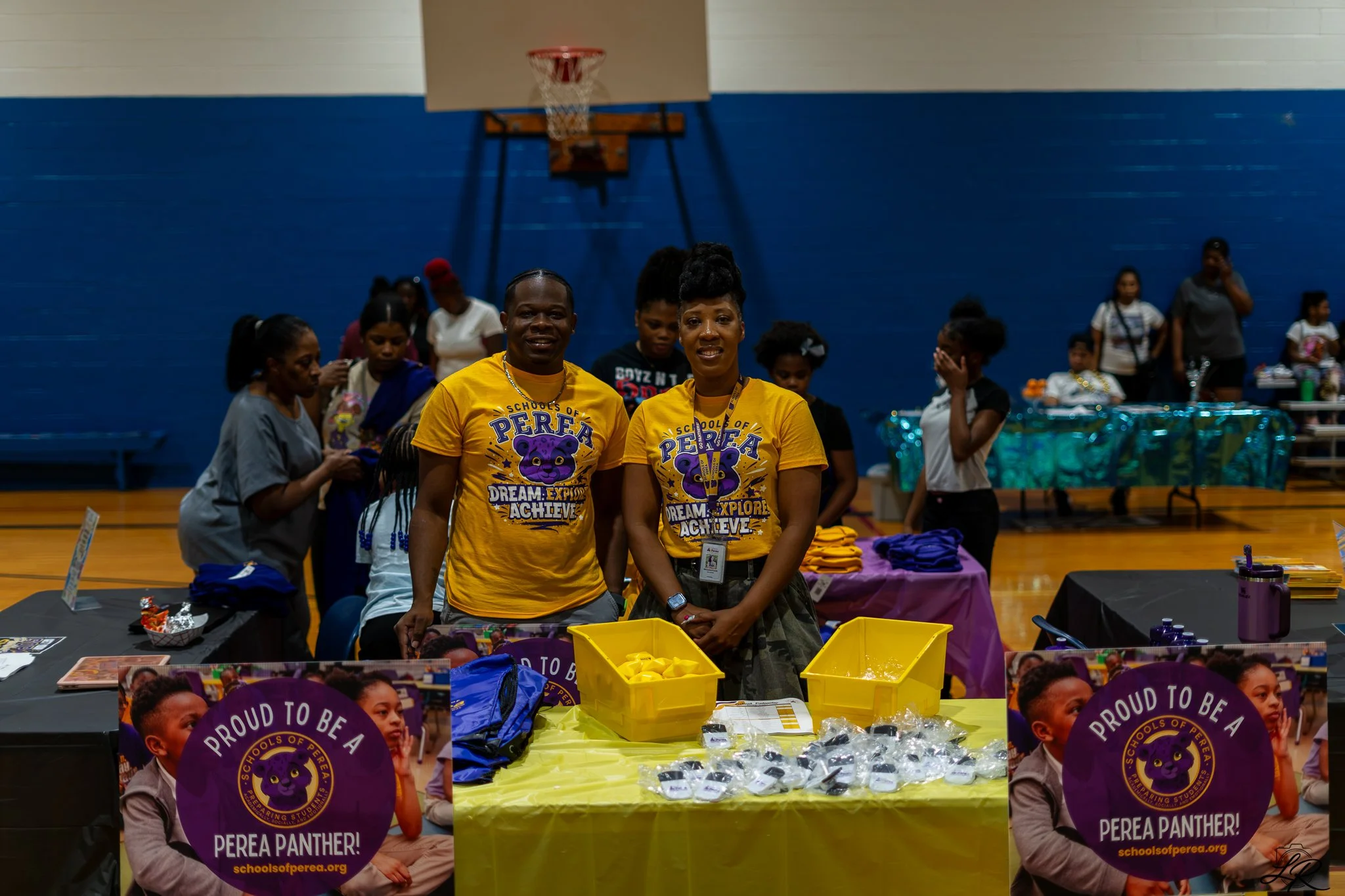 Indoor event at a school gymnasium with two women in yellow shirts standing behind a table with various items, surrounded by children and other adults, some holding signs that say "Proud to be a Perea Panther" with a panther mascot logo.