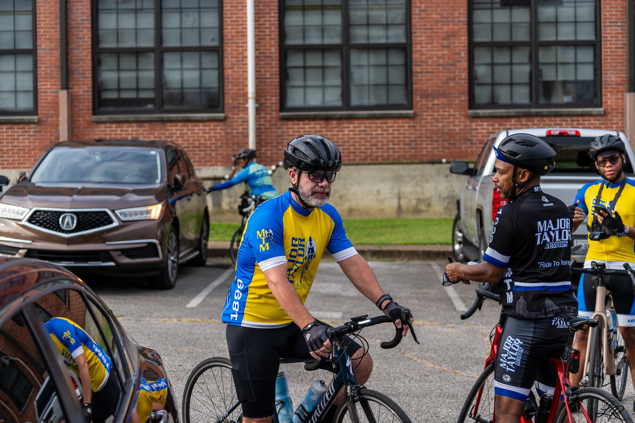 Group of cyclists in jerseys and helmets standing next to parked cars outside a brick building.