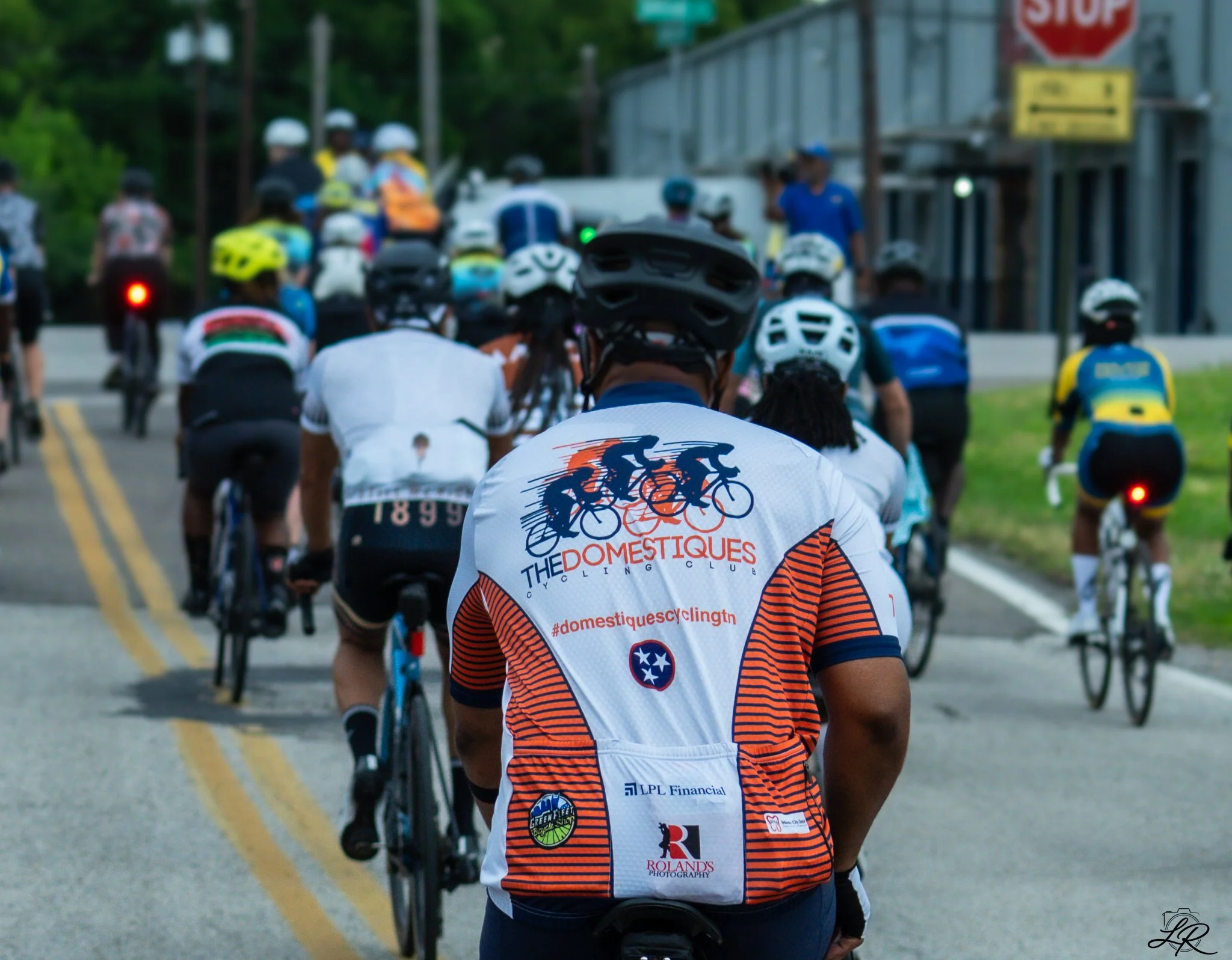 A group of cyclists riding on a city street, wearing helmets and athletic clothing, with a focus on a cyclist in the foreground wearing a jersey with various sponsors and logos.
