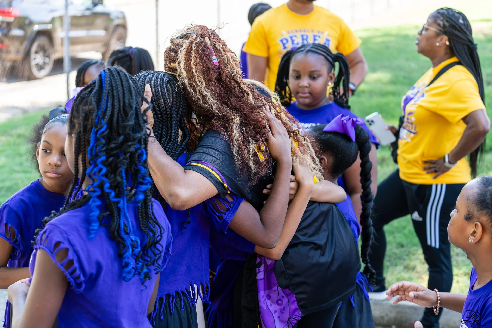 A group of women and children hugging and talking outdoors during daytime. Some are wearing purple shirts with cut-out sleeves, and yellow shirts with the word 'Perse' visible. The background shows a grassy area and a chain-link fence.