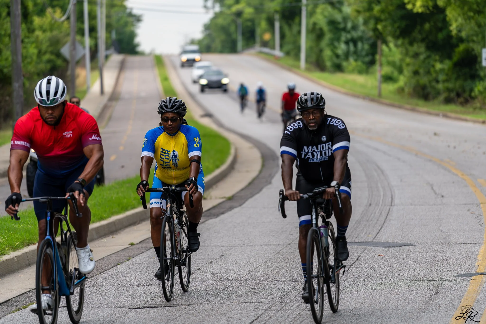 Group of four cyclists riding on a paved road with cars in the background, surrounded by green trees and grass.