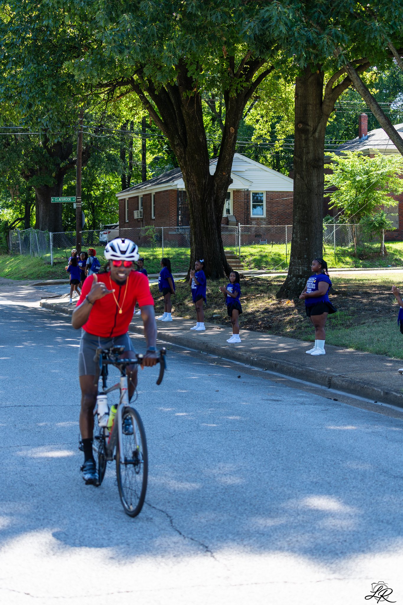 A cyclist in a red shirt and helmet riding on a street with young girls standing on the sidewalk watching. The background has trees, houses, and power lines.