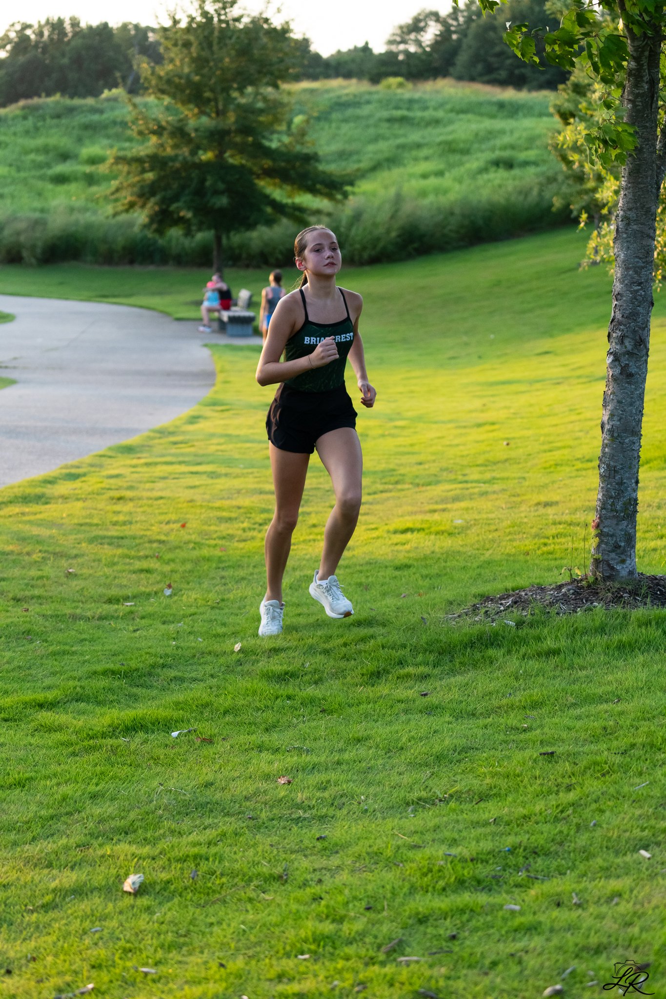 A young girl jogging on a park pathway with green grass and trees in the background. Two women are sitting on a bench further down the path, one wearing a dress and the other in sportswear, in a scenic outdoor setting during late afternoon or early e