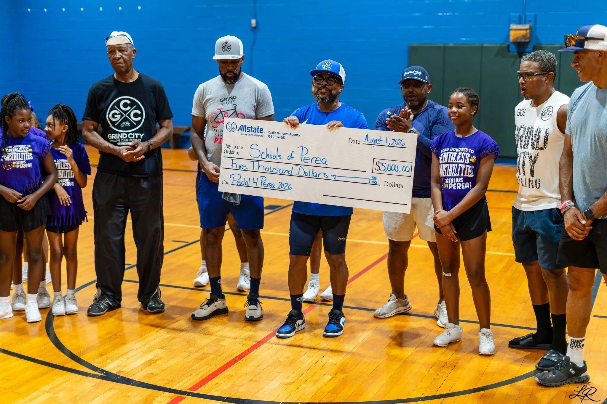 A group of people, including children and adults, standing on a gymnasium court during a check presentation. The adult man in the center holds a large check for $5,000 made out to Schools of Perea for Pedal 4 Perea 2026. The people are wearing athlet